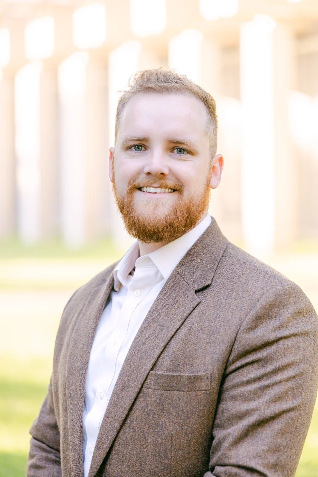 A man with a reddish beard, wearing a brown blazer and white shirt, smiling outdoors with a blurred background of vertical structures and sunlight.