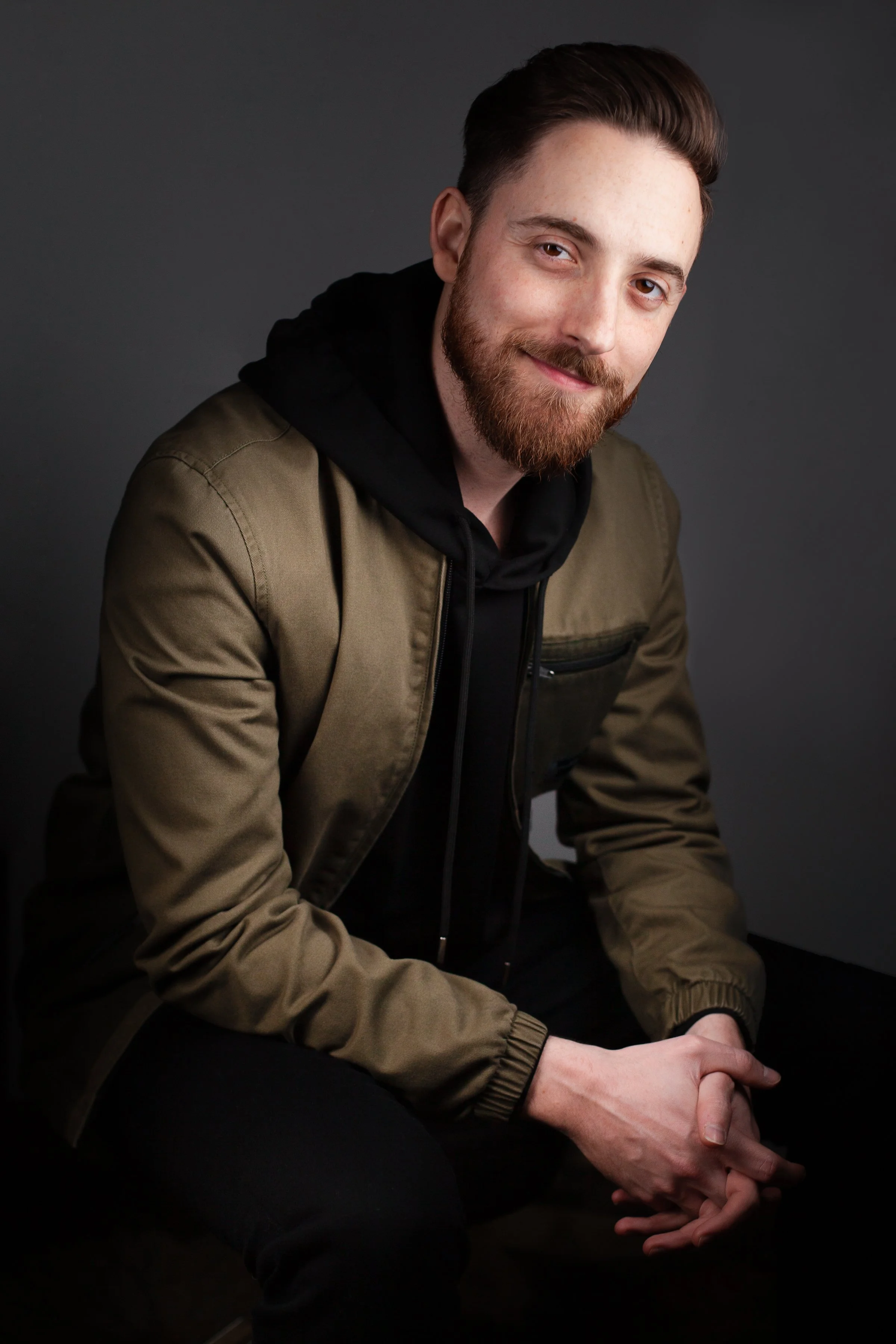 Drew Feldman, APMA®. A man with a well-groomed beard and styled hair, wearing a black hoodie and olive green jacket, sitting against a dark background, smiling gently and looking at the camera.