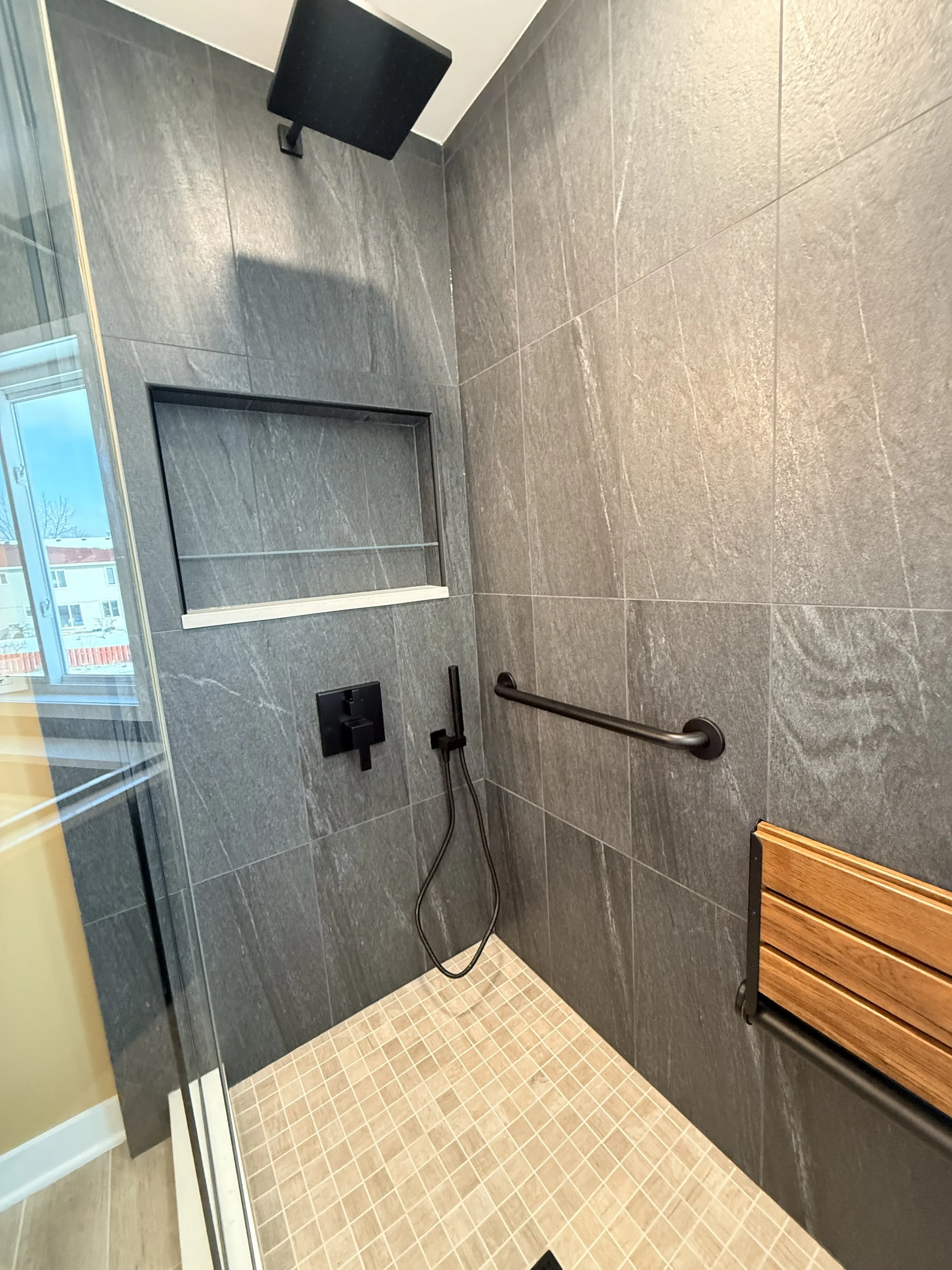 Luxury bathroom remodel walk-in shower with large gray wall tile, black rainfall shower head, built-in niche, safety grab bar, and mosaic tile shower floor, Wedi shower base
