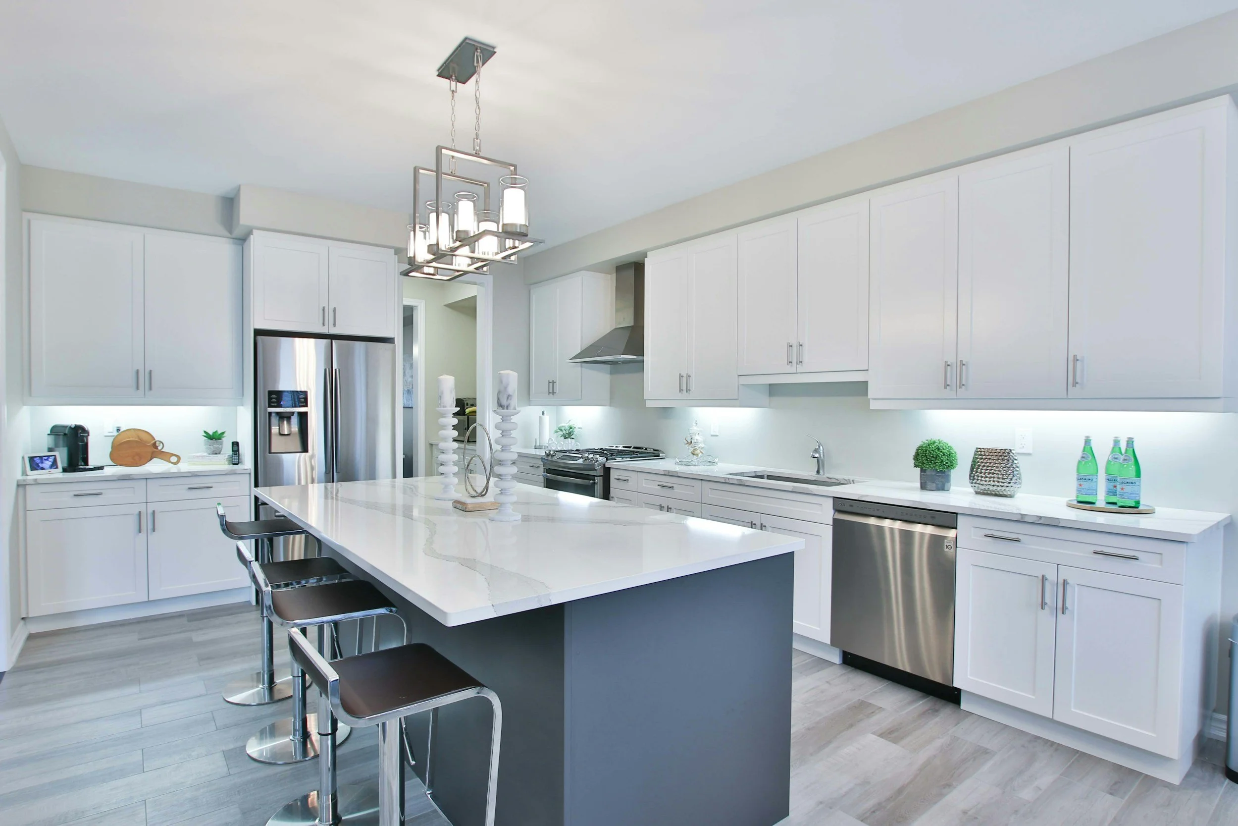 Modern kitchen with white cabinets, a large island with a white marble countertop, gray base, three black and chrome barstools, stainless steel appliances including a refrigerator, oven, and dishwasher, decorative candles on the island, potted plants, water bottles, and a sleek chandelier hanging from the ceiling.