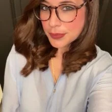 A woman with long brown hair, wearing glasses and a white top, looking at the camera.