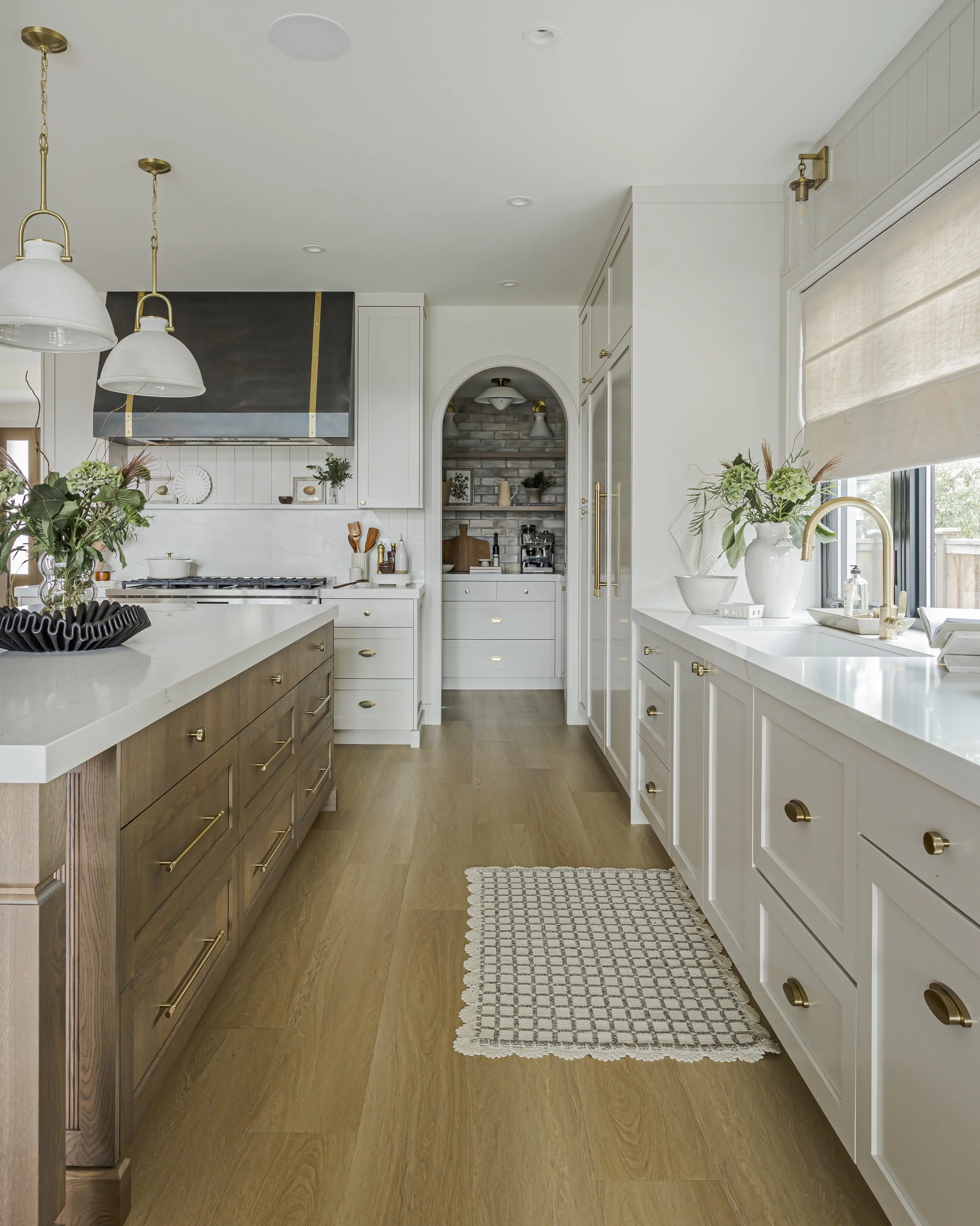 Kitchen with a dark wood island, white perimeter cabinetry, brass fixtures, and a walk-in pantry visible through an arched doorway