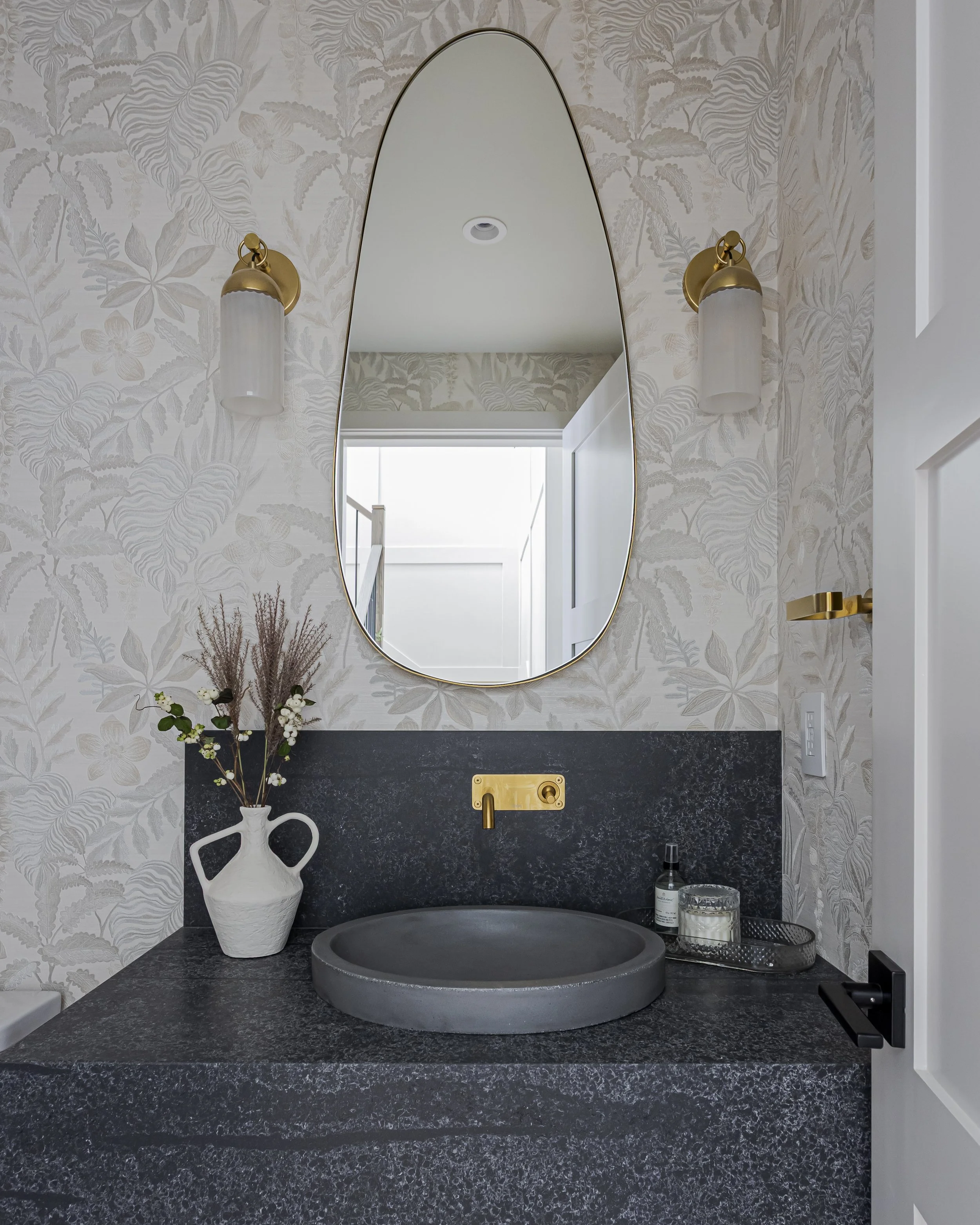 Powder room with patterned wallpaper, a black stone vessel sink, brass fixtures, and an oval mirror