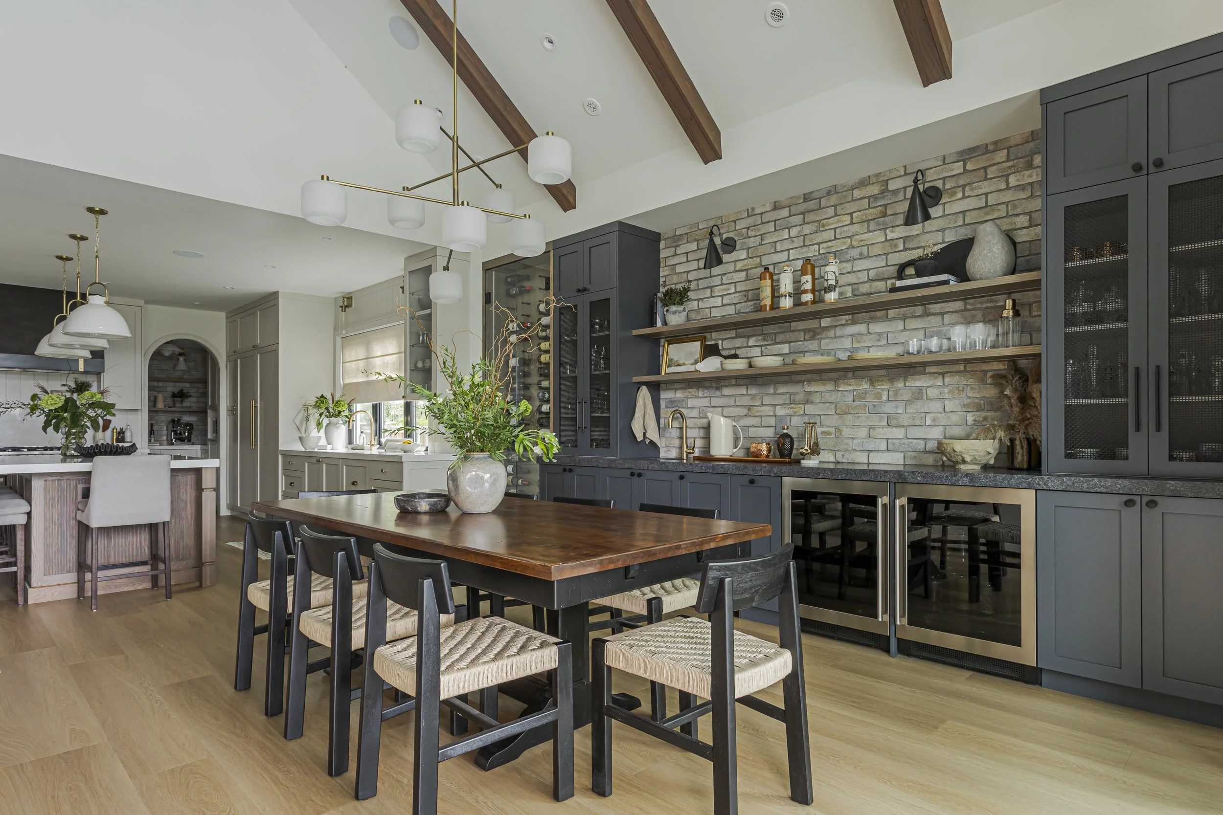 Open-concept kitchen and dining area with a wood dining table, black chairs, dark gray cabinetry, brick backsplash with open shelving, and a built-in wine fridge.