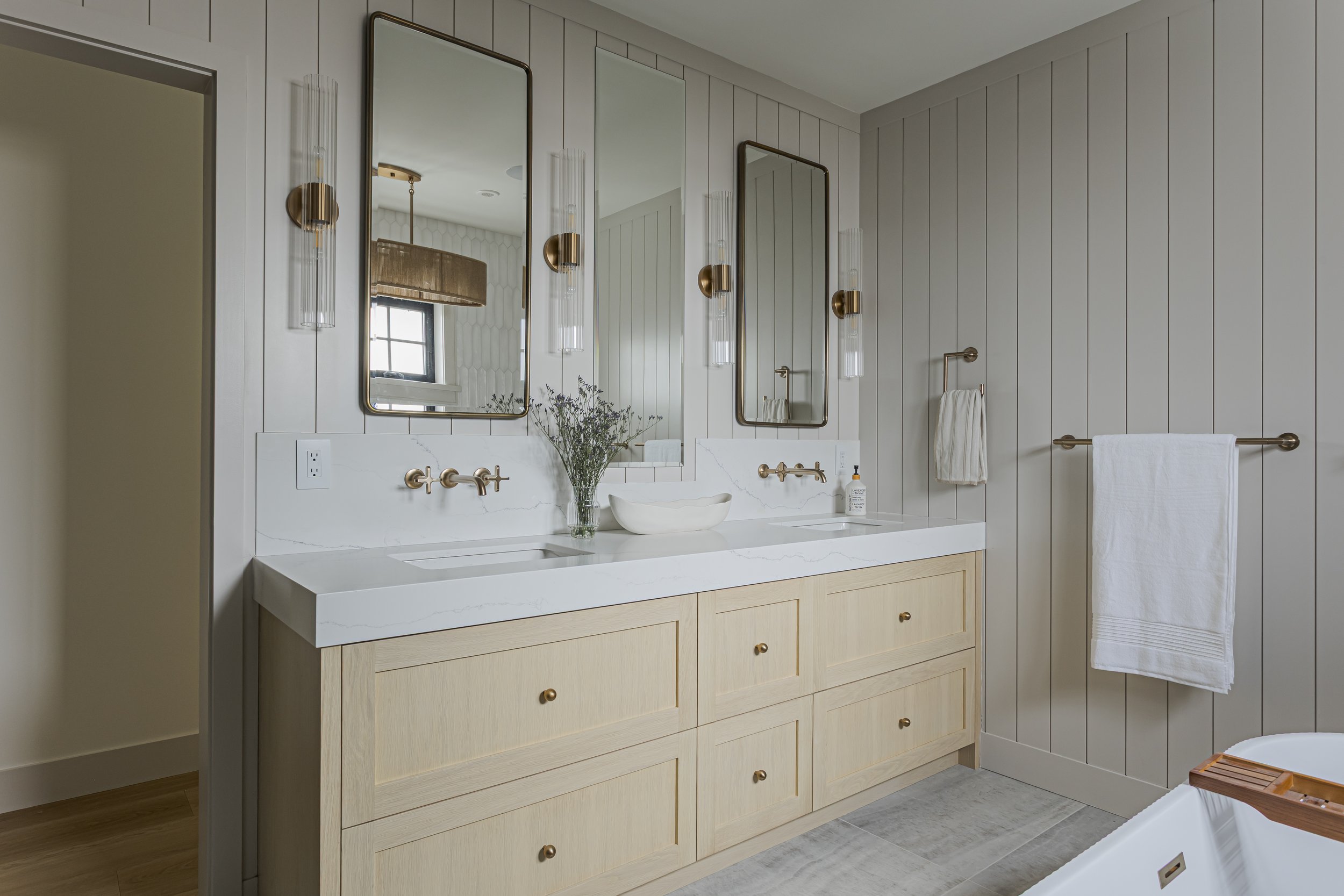 Primary bathroom with a light wood double vanity, brass hardware, and framed mirrors with sconce lighting.