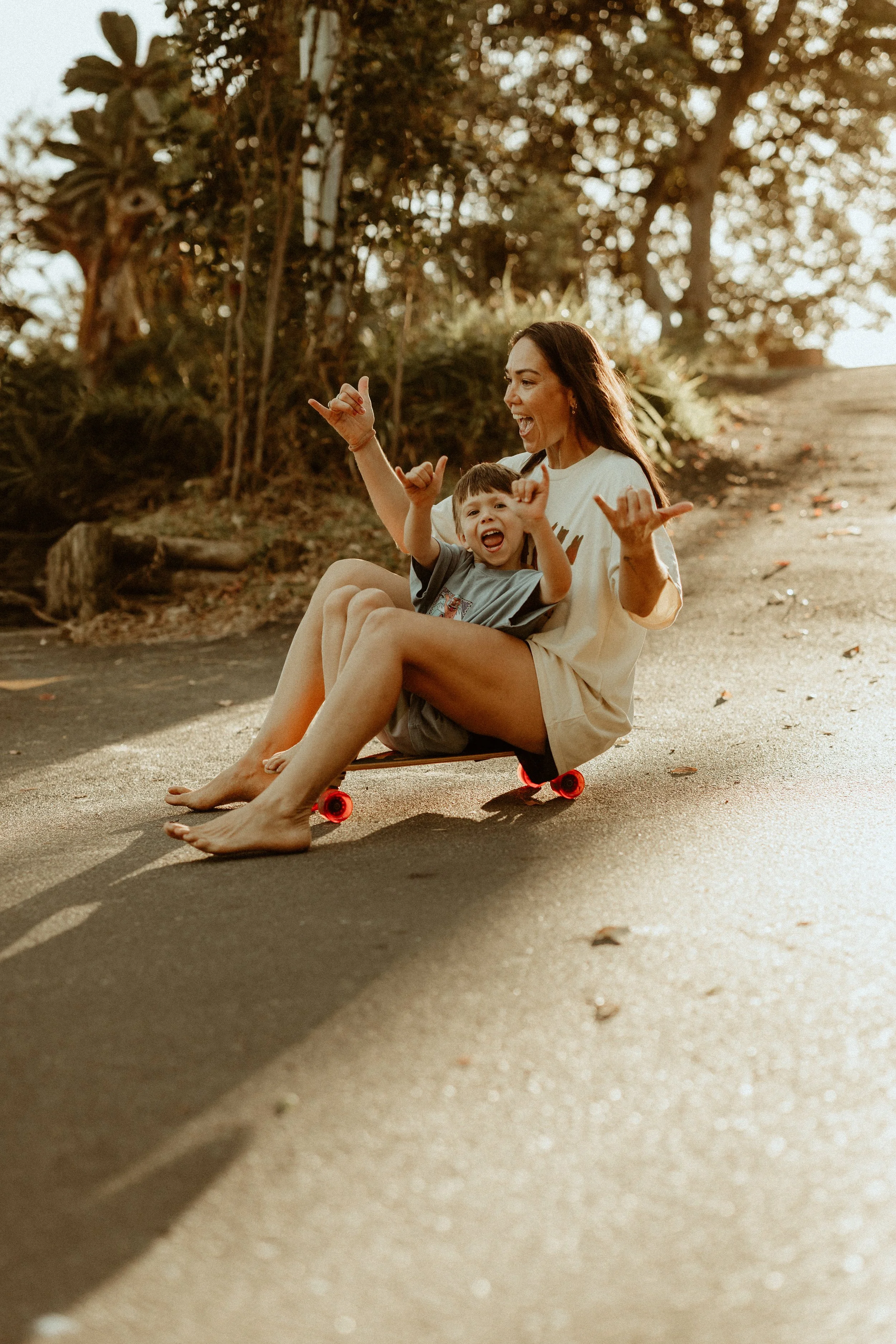 A woman and a young boy riding a skateboard together outdoors on a sunny day, smiling and making playful gestures.