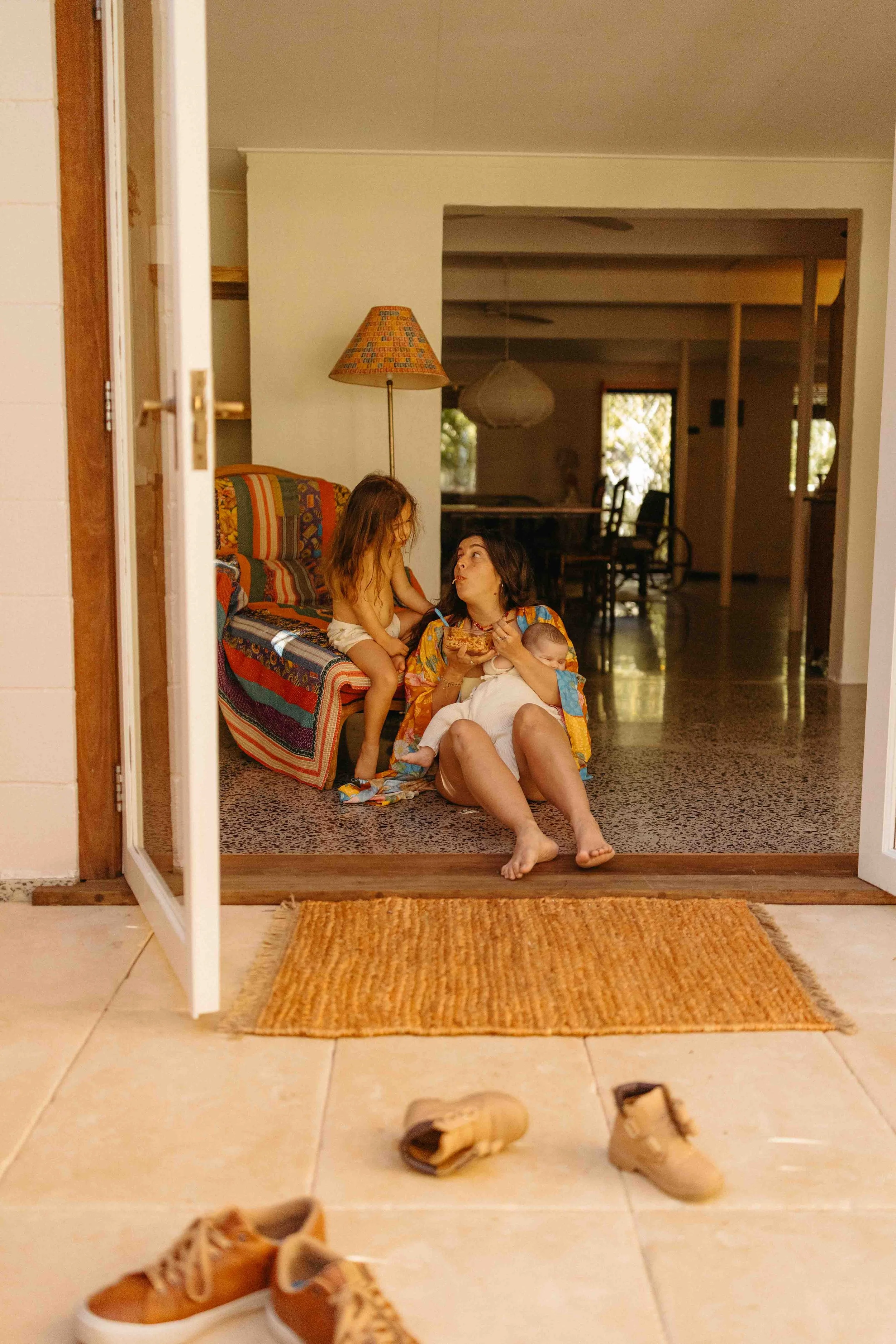 A mother with a baby in her arms is sitting on the floor, just inside her patio door eating spaghetti out of a glass container. Her toddler daughter is right next to her sitting on an armchair as they look at each other in a sweet affectionate way.