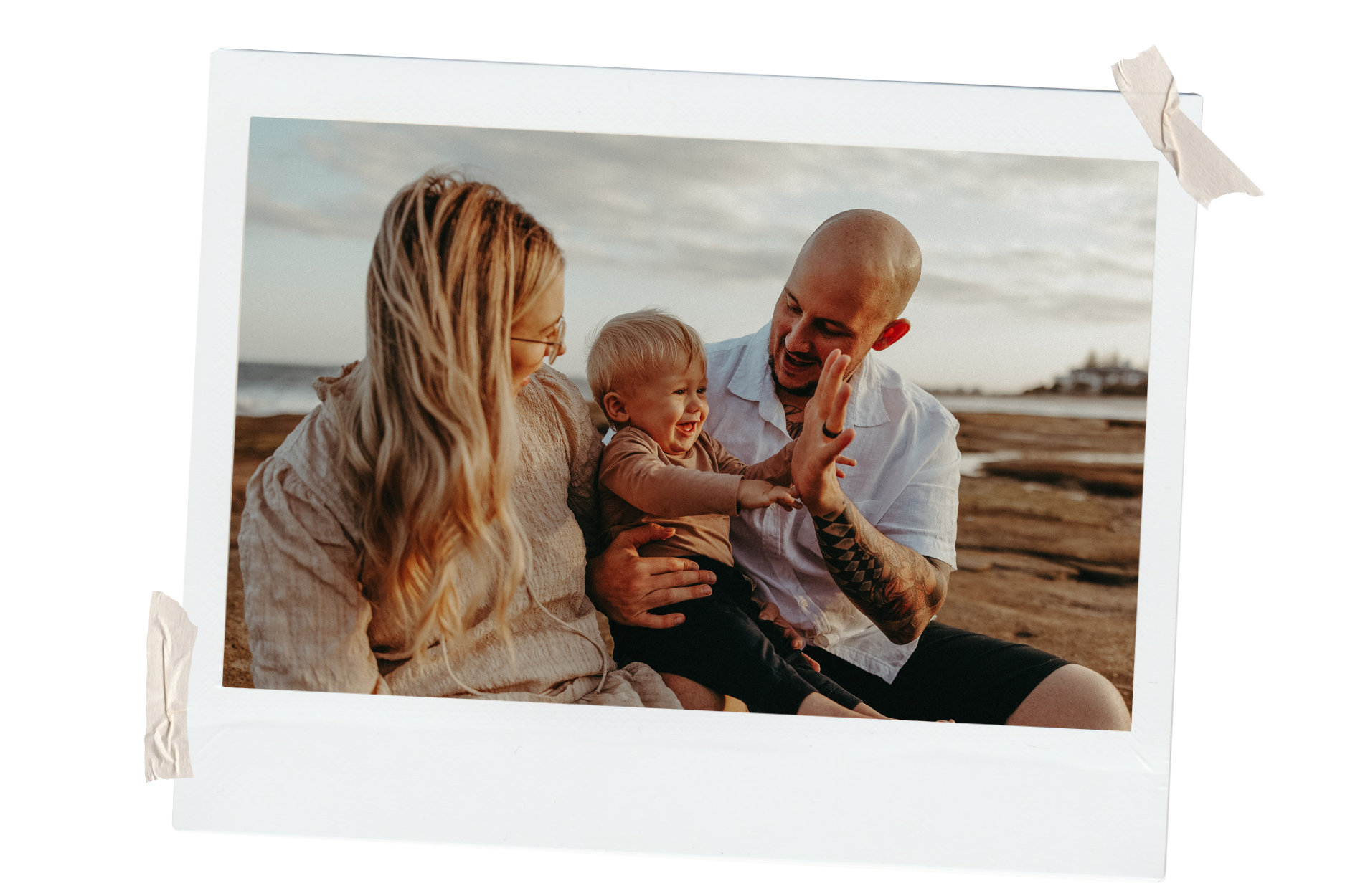 A family of three sitting outdoors on a beach during sunset, smiling and engaging with each other. The father is bald with tattoos, holding his young son who is giving a high five to the mother. The mother has long blonde hair and glasses.