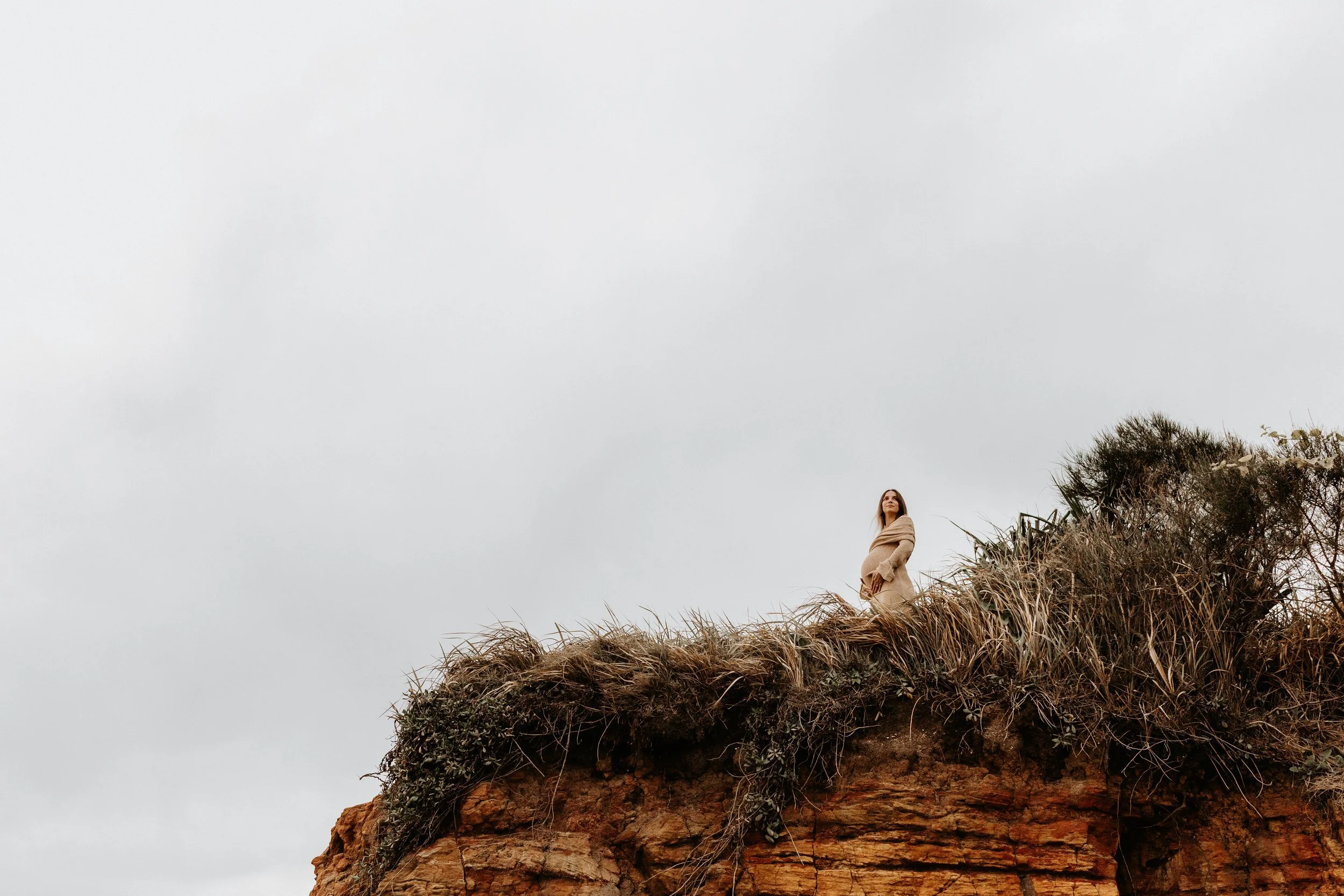 A woman standing on the edge of a rocky cliff covered with grass and shrubs, looking outward with an overcast sky in the background.