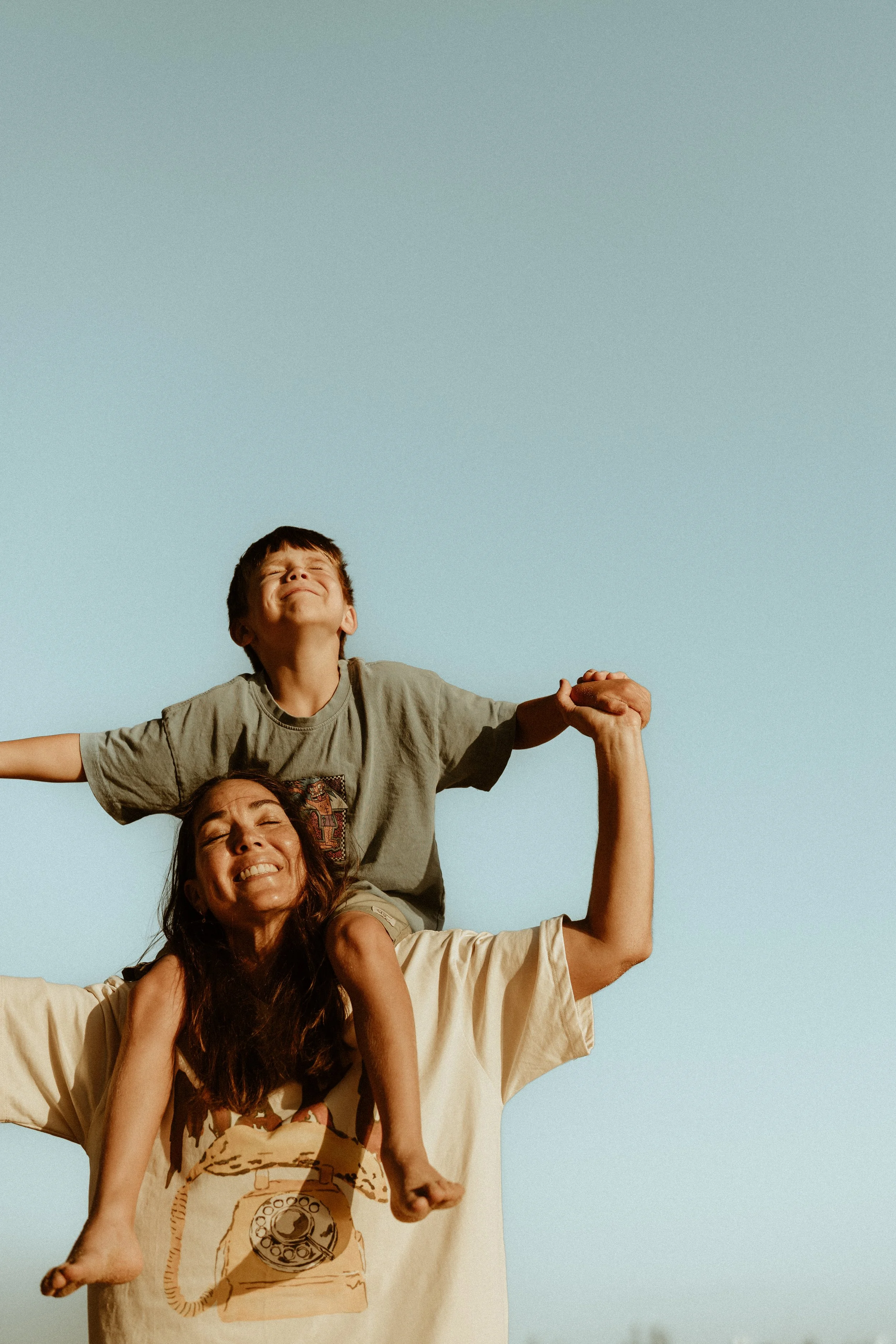 A woman and a young boy are playing outdoors under a clear blue sky. The woman is smiling and lifting the boy onto her shoulders, who has his arms outstretched and eyes closed in joy.