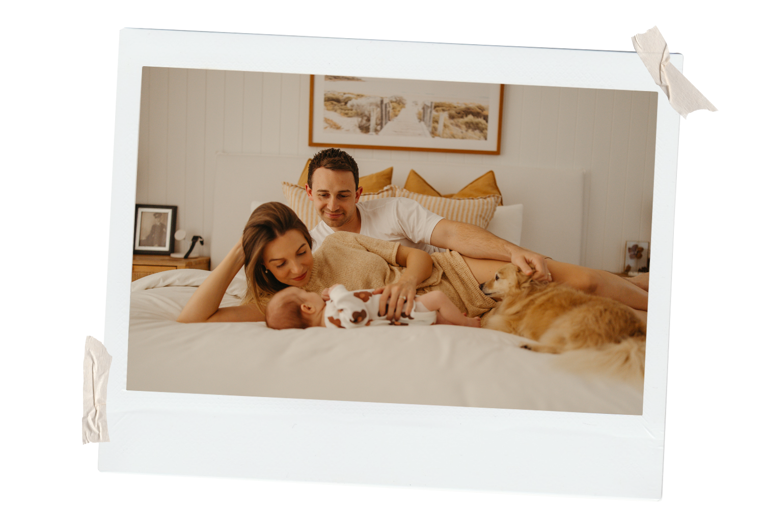 A family with a mother, father, and newborn baby lying on a bed with a dog in a cozy, well-lit bedroom.