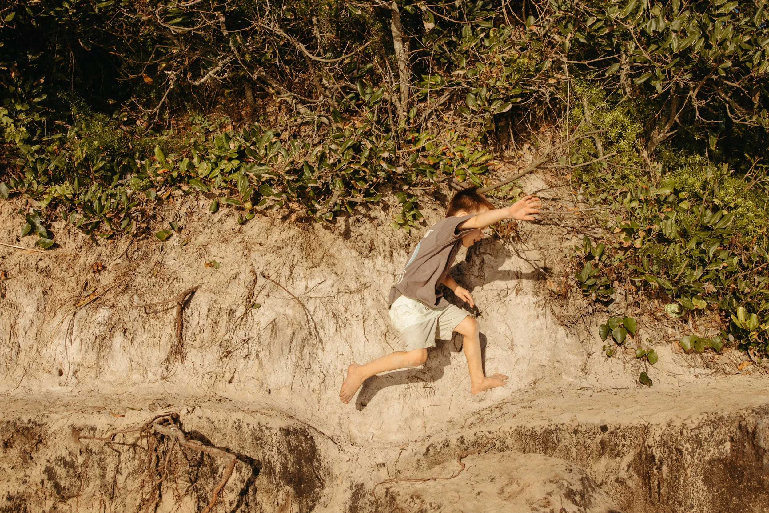 A boy in a brown shirt and beige shorts running barefoot on sandy ground near green bushes and trees.