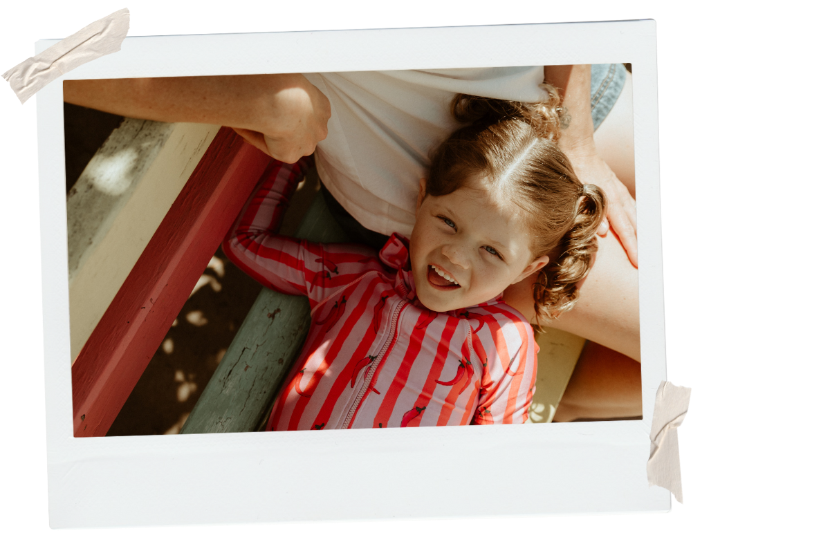 A young girl with curly hair, wearing a red and white striped jacket with flamingo patterns, smiling and lying on a playground structure with red and green bars.
