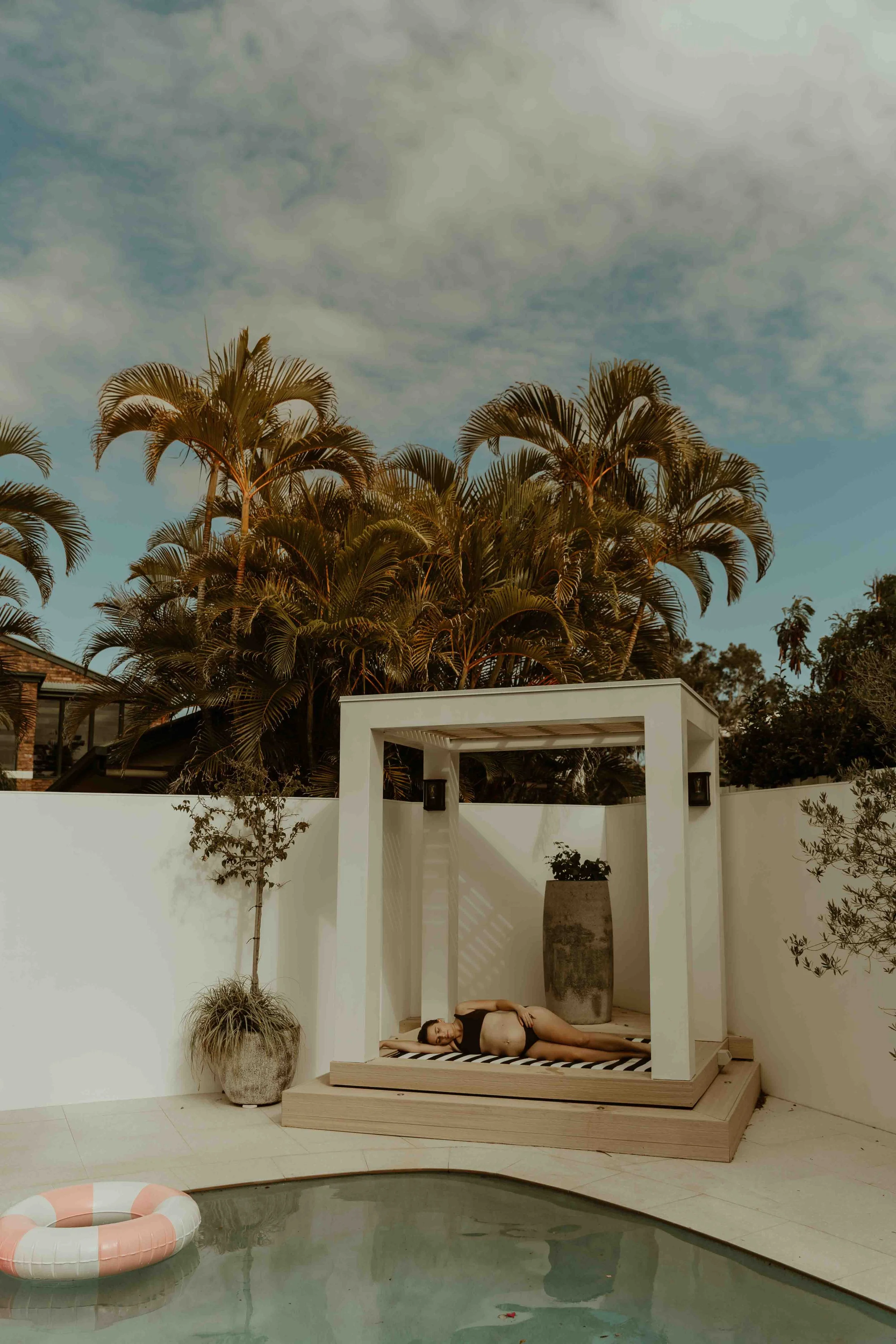 A pregnant woman is lying underneath a poolside pagoda on her side. There are tall palmtrees and blue sky in the background and a pink and white inflatable pool ring floating in the foreground.