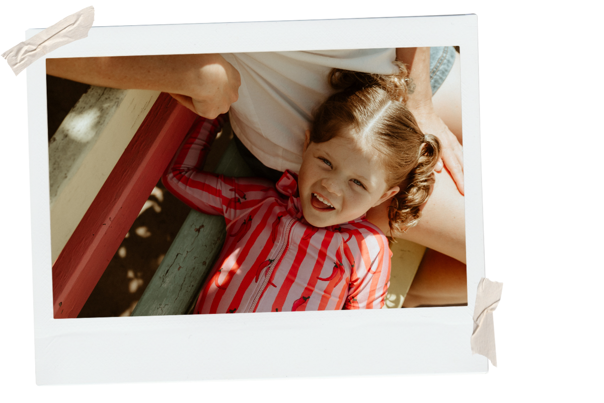 A young girl with curly brown hair lying on a playground surface, smiling and looking up, wearing a red and white striped jacket with flamingo prints, partially covered by an adult who is sitting beside her.