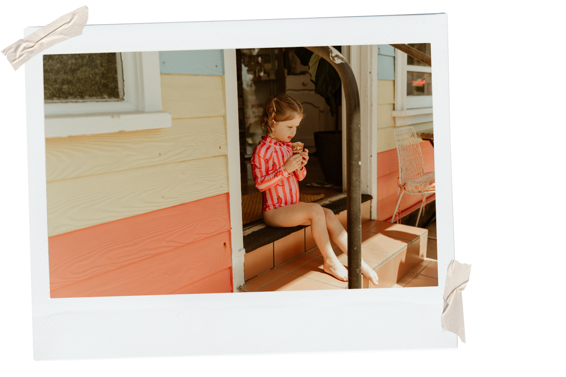A young girl in a red and white striped shirt is sitting on a porch step, eating a cupcake. The porch has a colorful wooden exterior with shades of cream and coral, and a white window and door in the background. There is a chair with an orange cushion to her right.