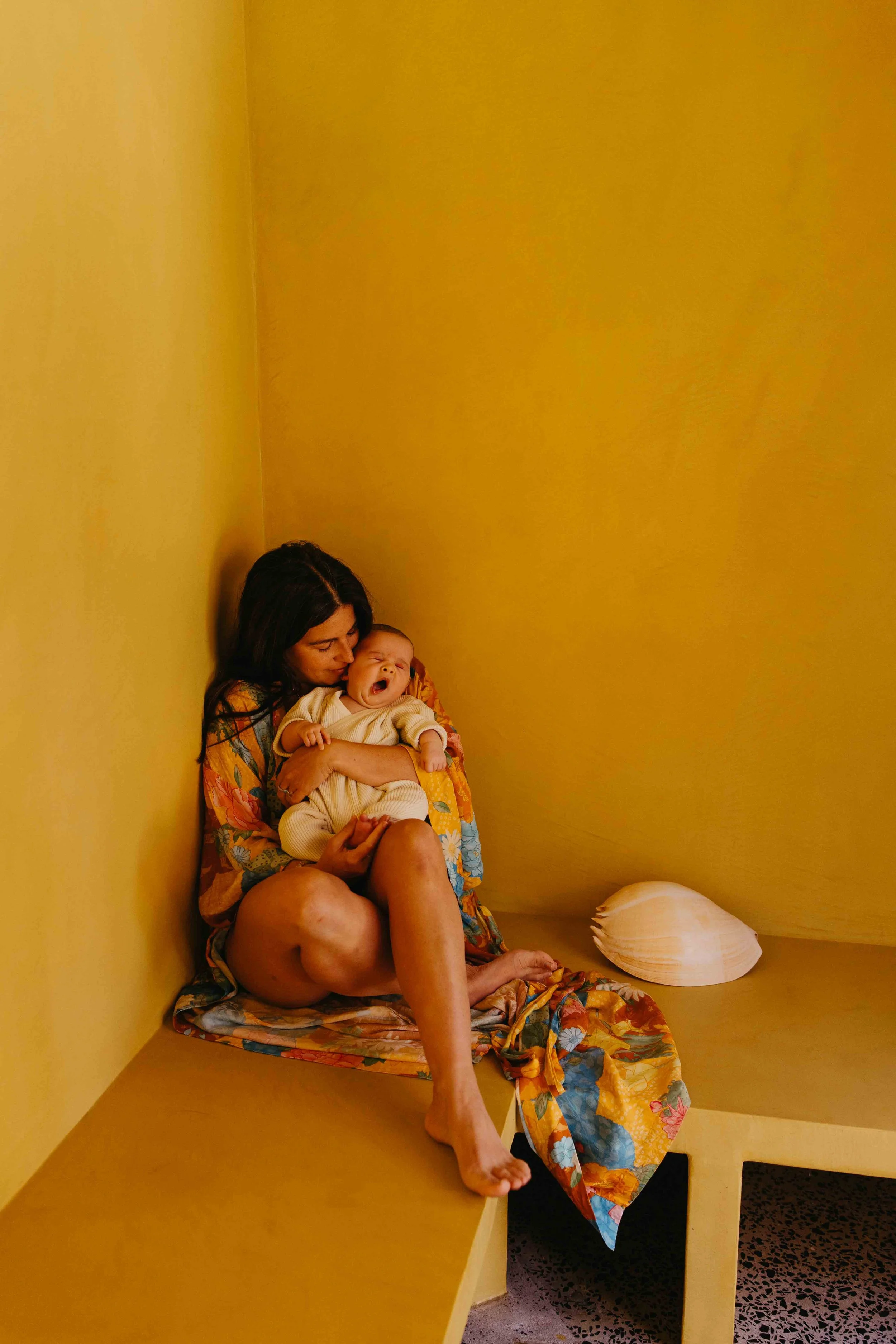 Mother sitting in the corner of a small, box-like space with bright orange yellow walls holding her baby in her arms. The baby is yawning while the mother leans in close, gently comforting and kissing baby's cheek.