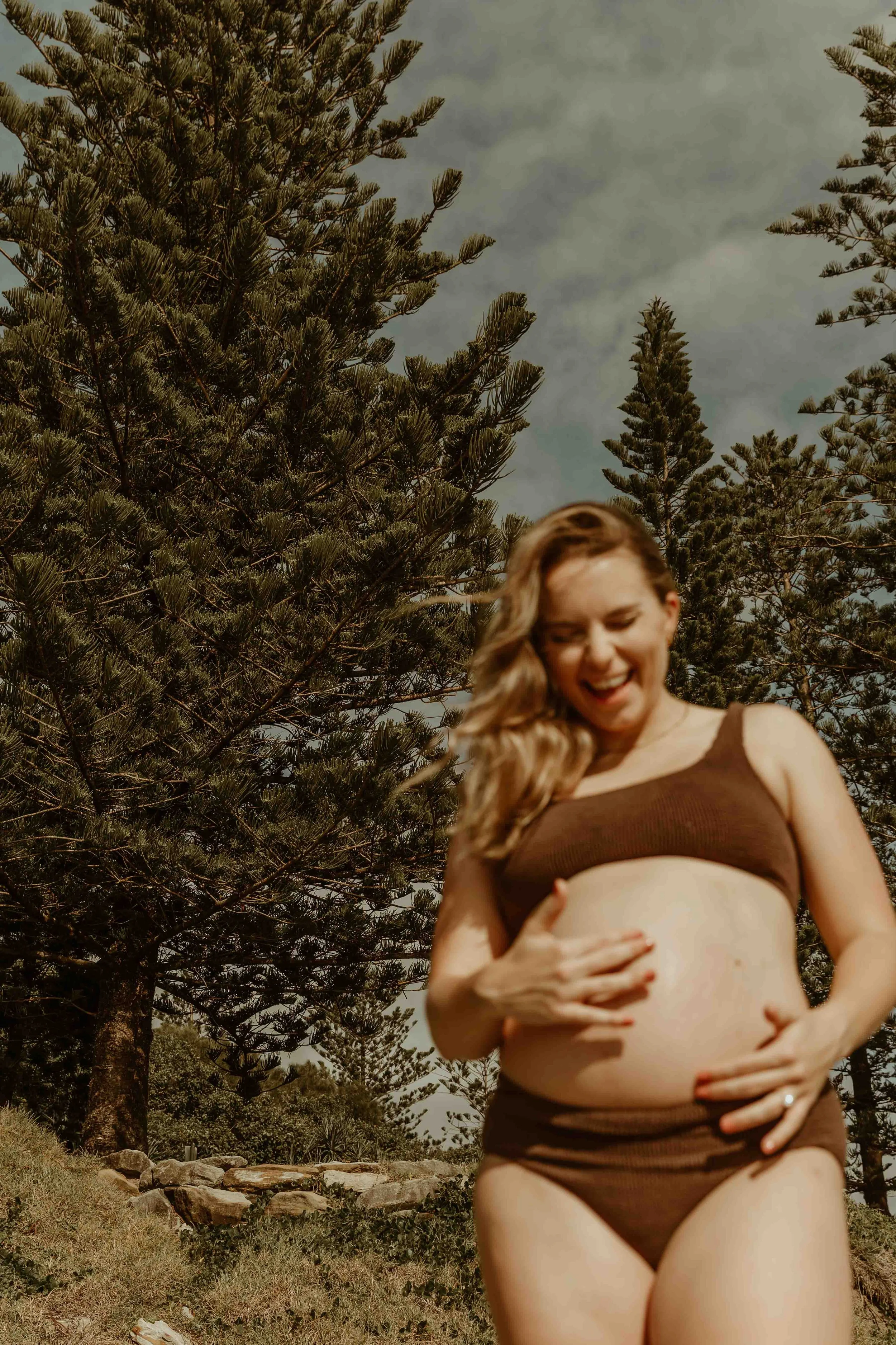 A pregnant woman tickling her belly button laughing in the foreground slightly out of focus with large, pine trees behind her under a clear blue sky.