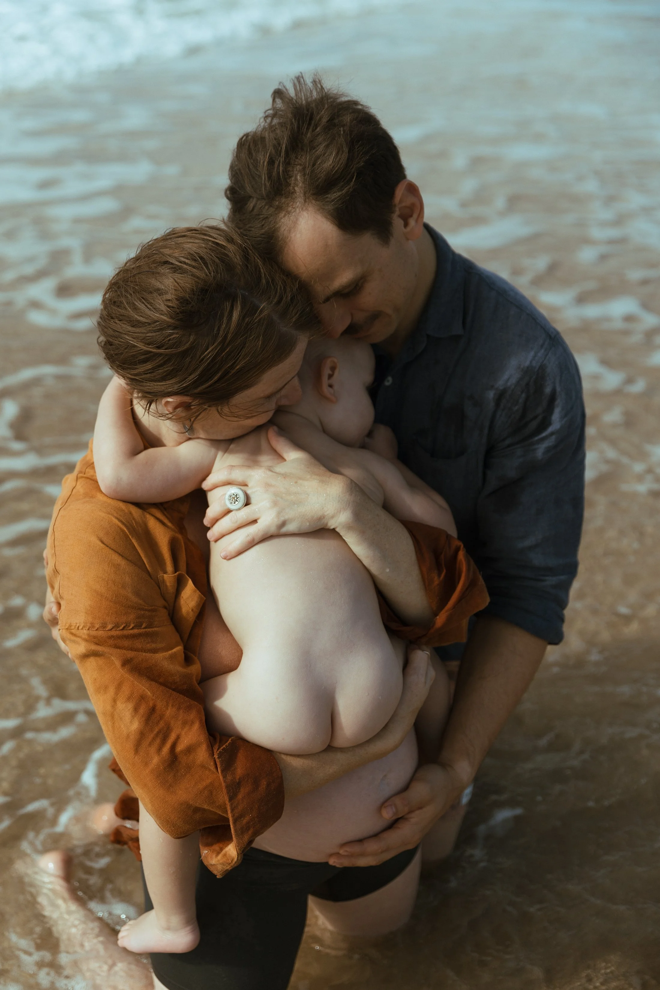 A family on the beach holding their baby and sharing an intimate moment as they stand in the water.
