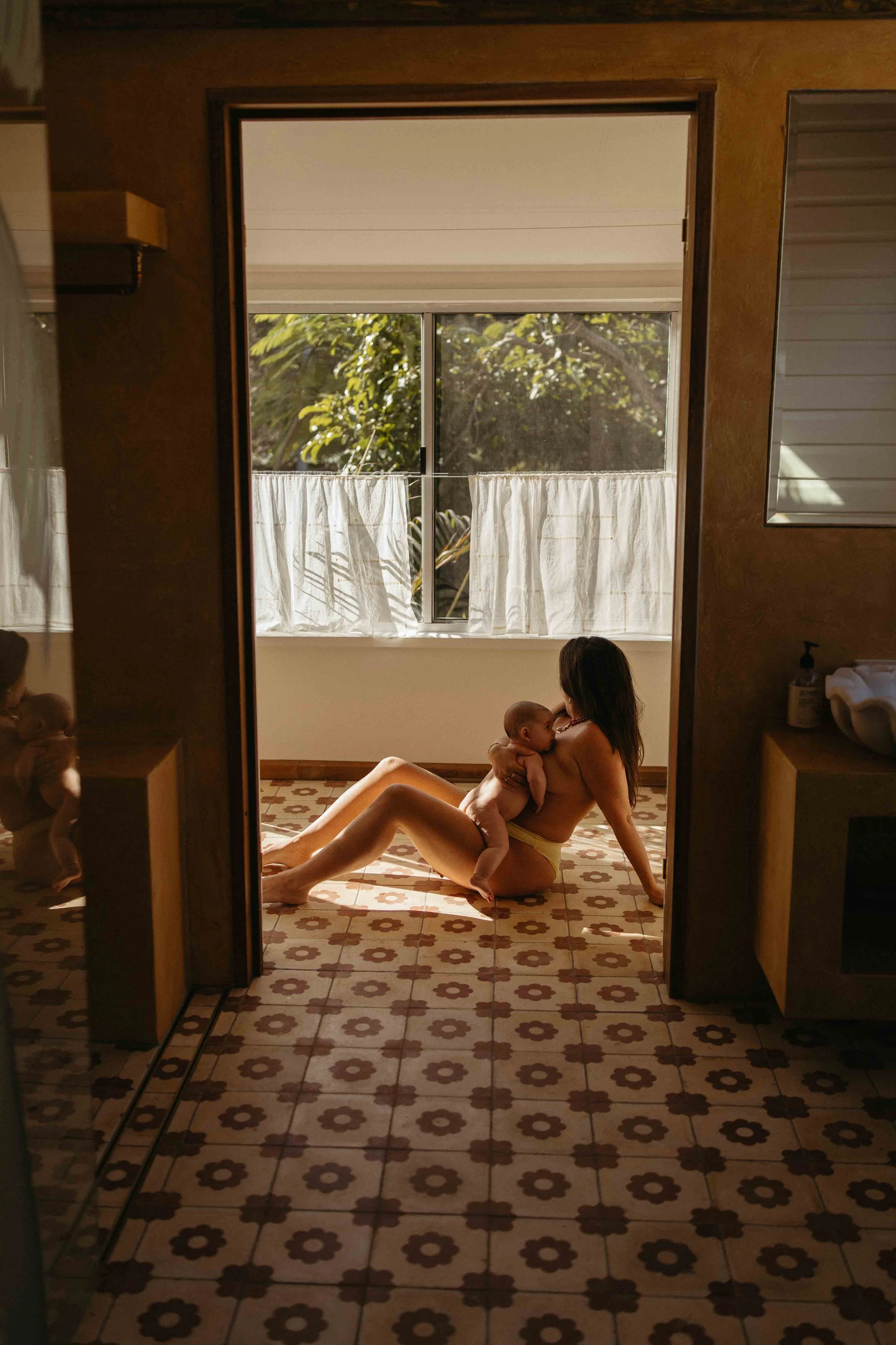Mother nursing 3 month old baby on floral mosaic tiled bathroom floor, basking in natural sunlight shining through window.