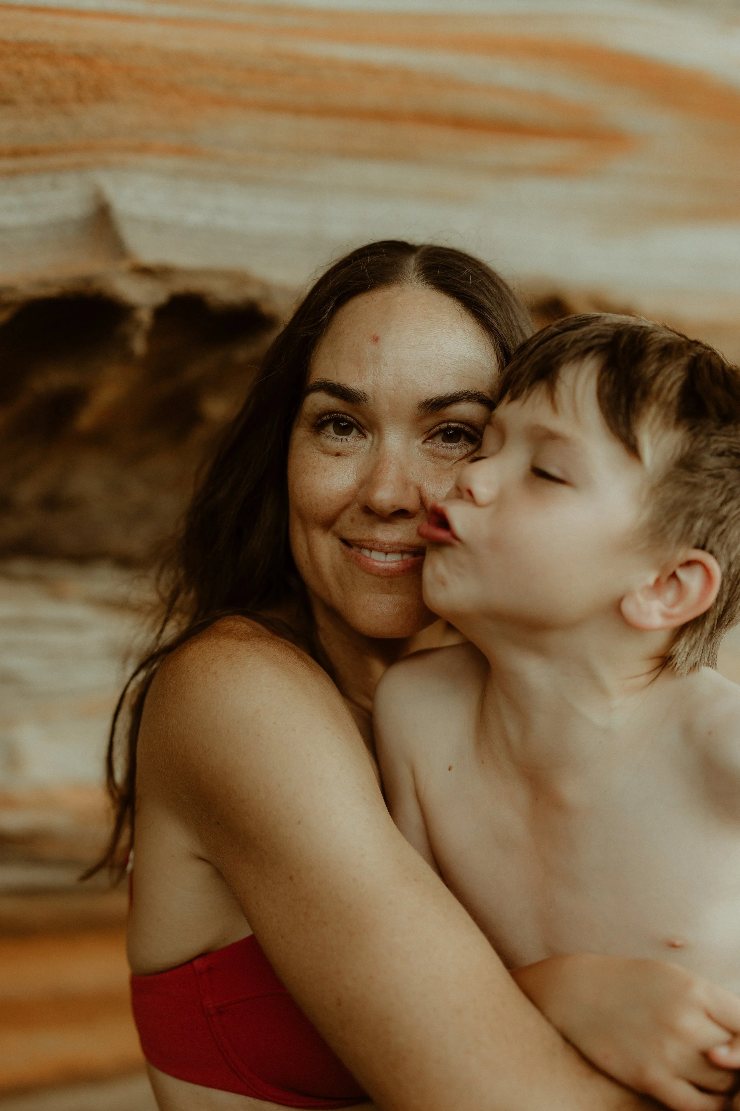 A woman with long dark hair smiling while holding a young boy with short light brown hair, who is kissing her cheek.