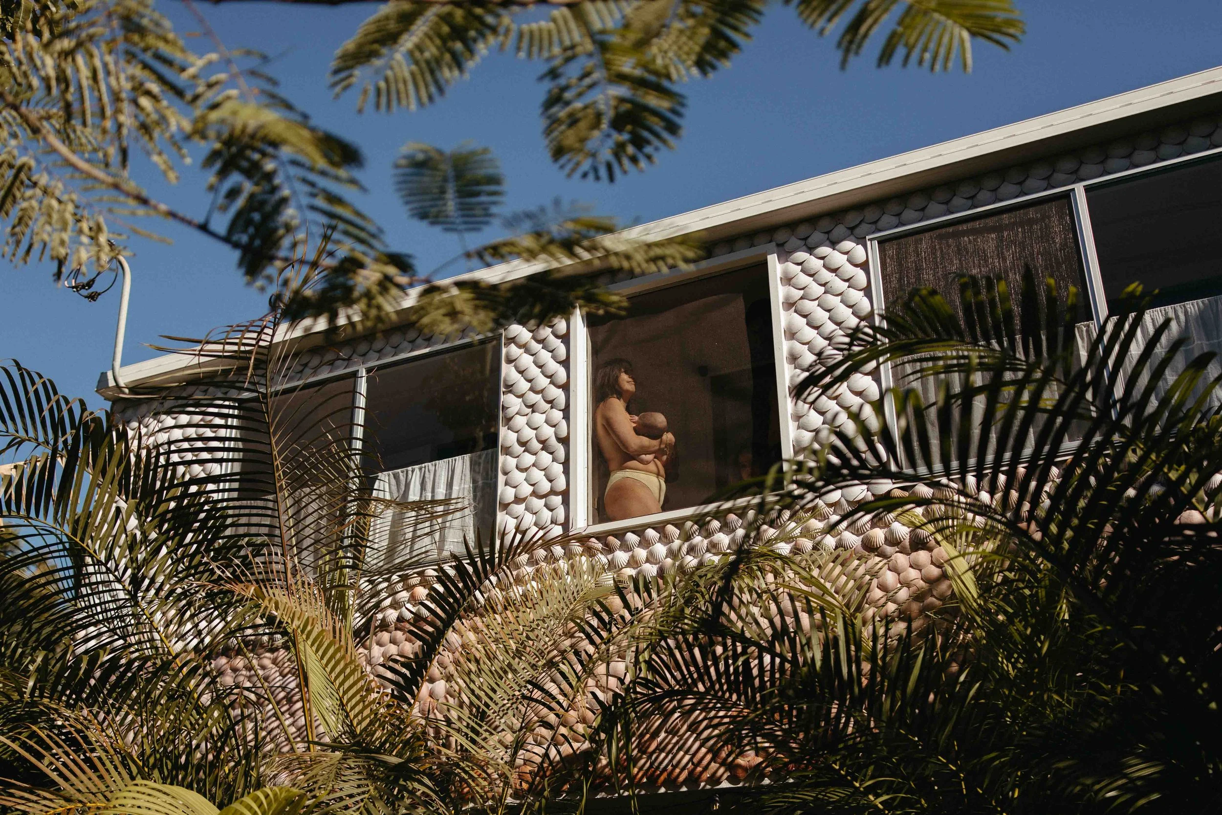 View of mother holding newborn through second floor window. The home is covered in giant clam shells and surrounded by lush, green tree foliage.