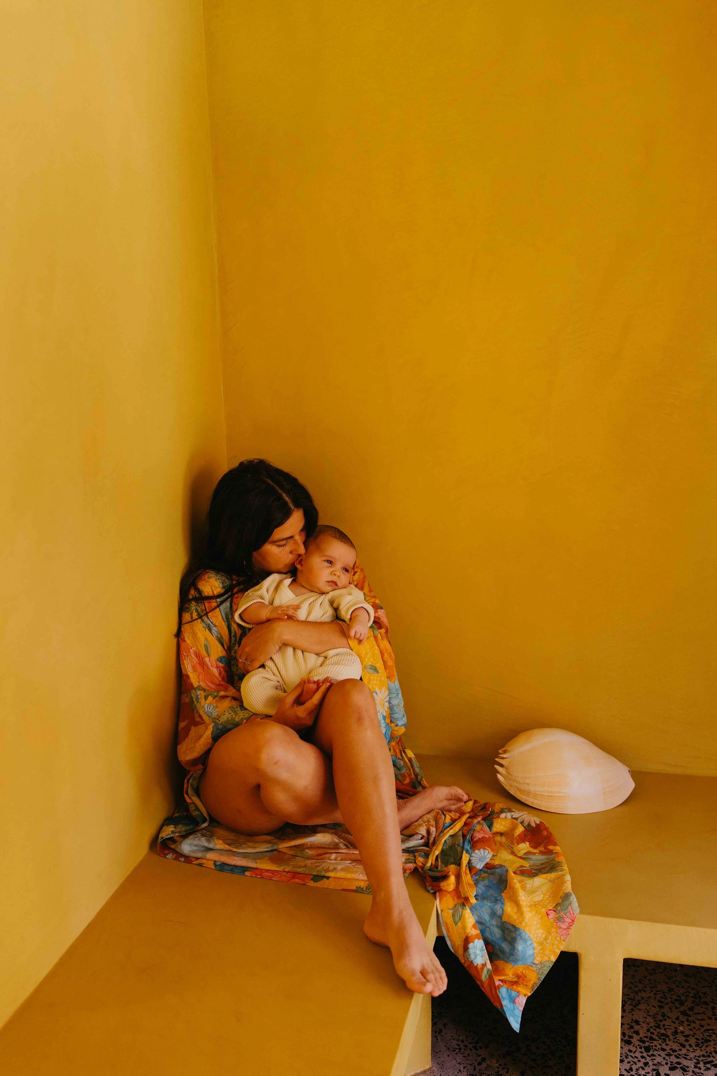 A mother seated in a corner cuddling her newborn baby, gently kissing her ear. The baby has wide eyes open staring off to the right.