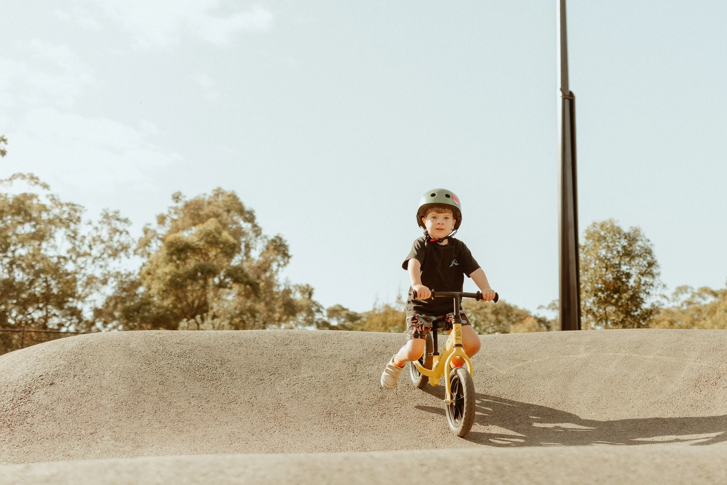 Young boy wearing a helmet riding a yellow balance bike on a dirt track in a park with trees in the background.