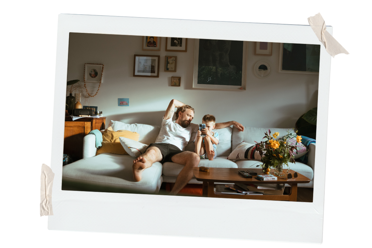 A man and a young boy sitting on a light-colored sofa in a living room, with the man leaning back and the boy holding a smartphone, with sunlight illuminating the room and framed pictures on the wall behind them.