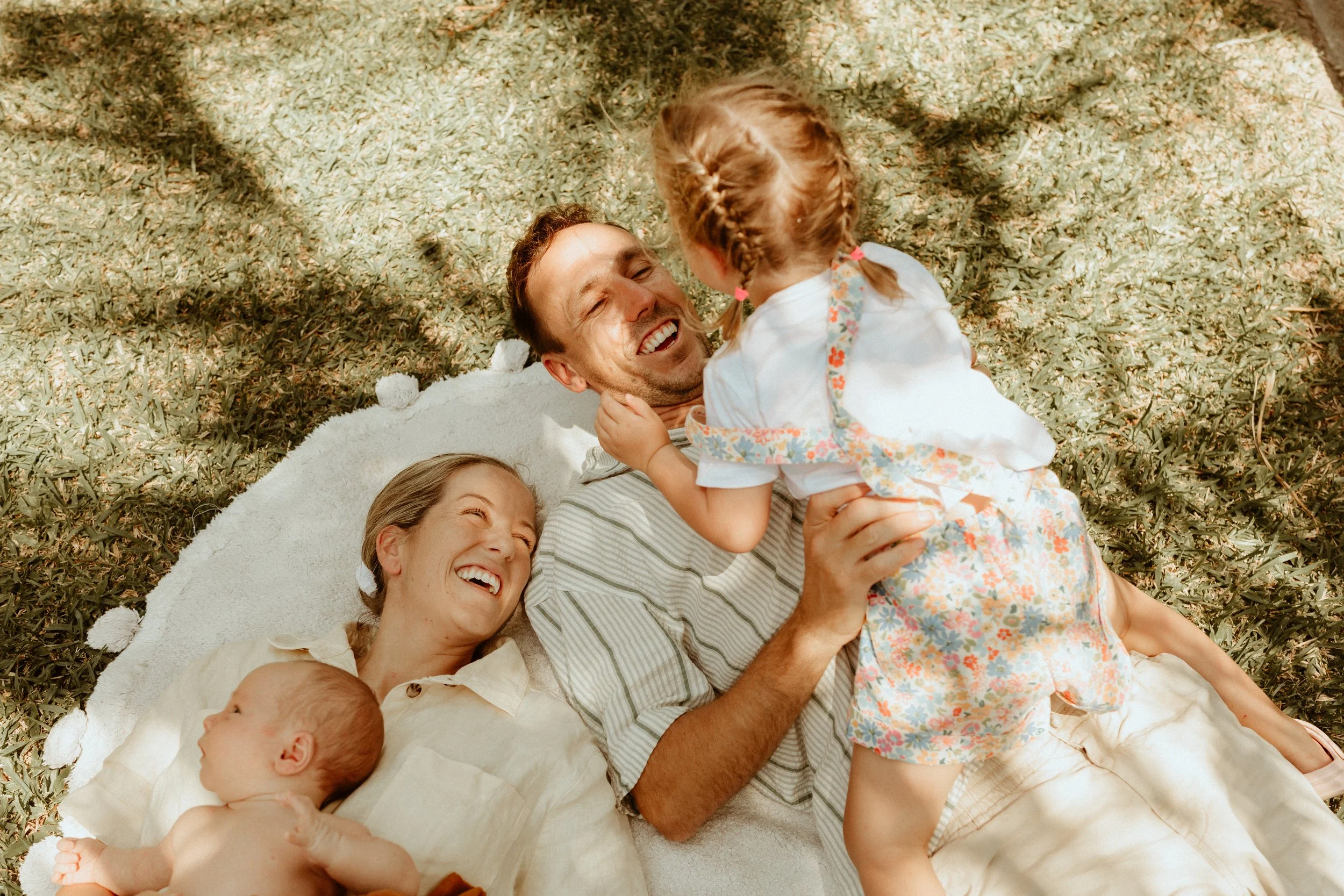 A family of four lying on a white blanket outdoors, with a tree overhead. The father is smiling and holding a young girl who is sitting on him, the mother is lying next to them and laughing, and a baby is lying beside the mother.