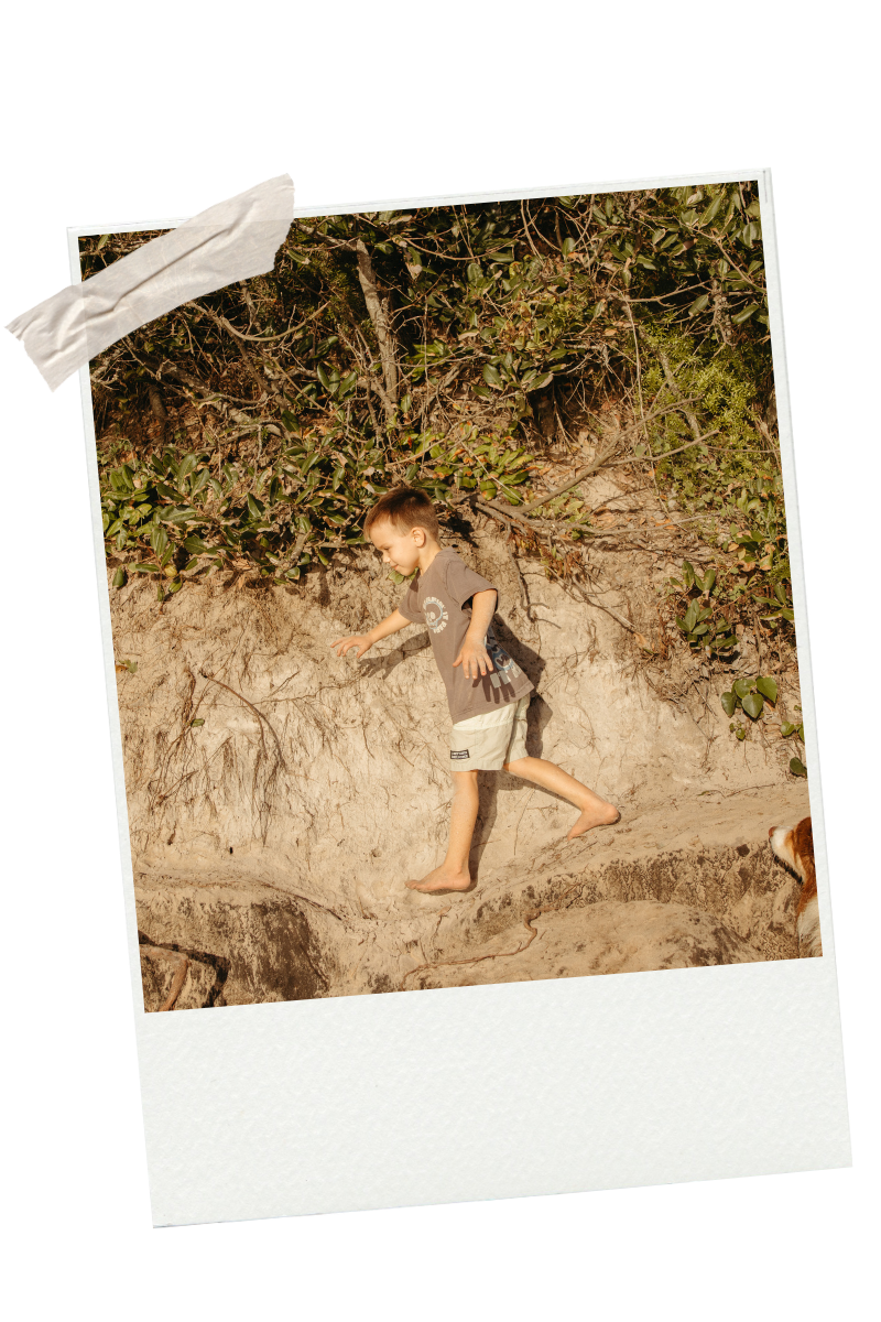 A young boy in a gray t-shirt and white shorts walking barefoot on a sandy and slightly rocky terrain outdoors, with green bushes and plants in the background.