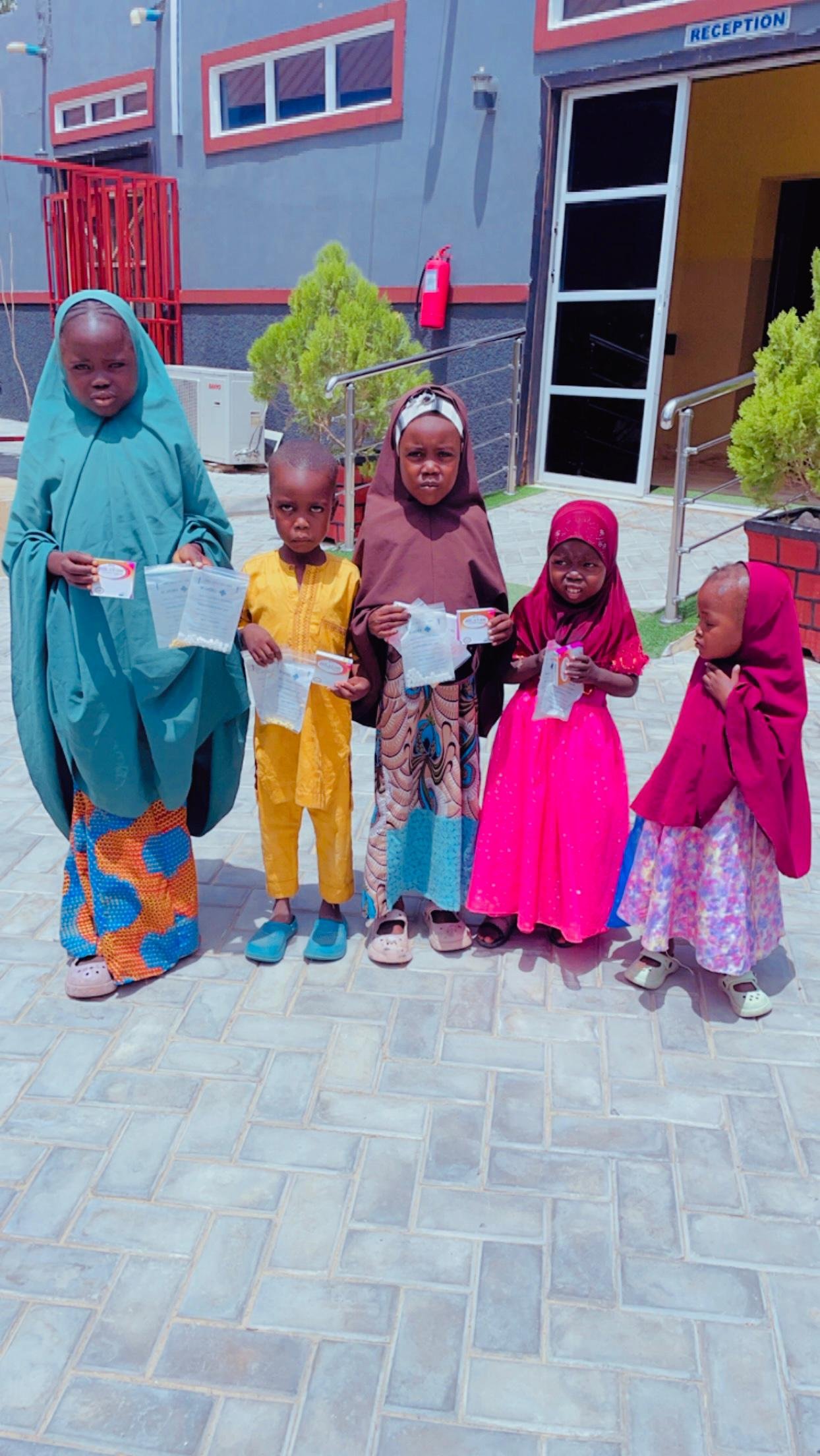 Five children standing outside a building holding papers and cards, wearing colorful traditional clothing and headscarves.