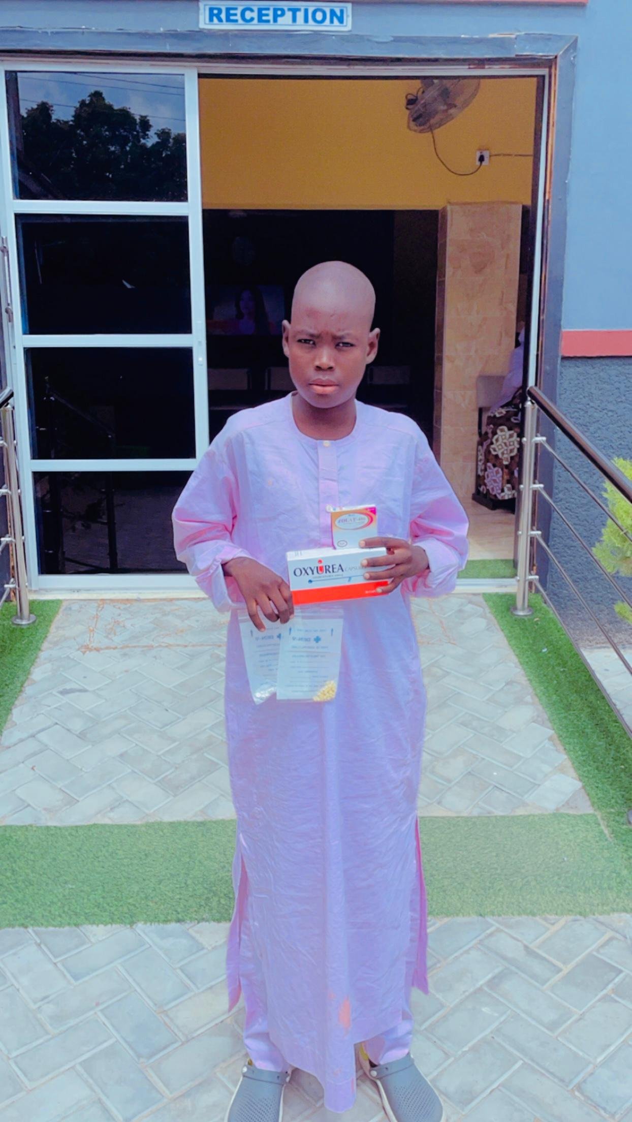 A young boy with a shaved head wearing a light pink traditional outfit standing outside a building labeled 'Reception'. He is holding boxes of medication, one labeled 'Oxyurea' and some documents, standing on a tiled pathway with artificial grass on 