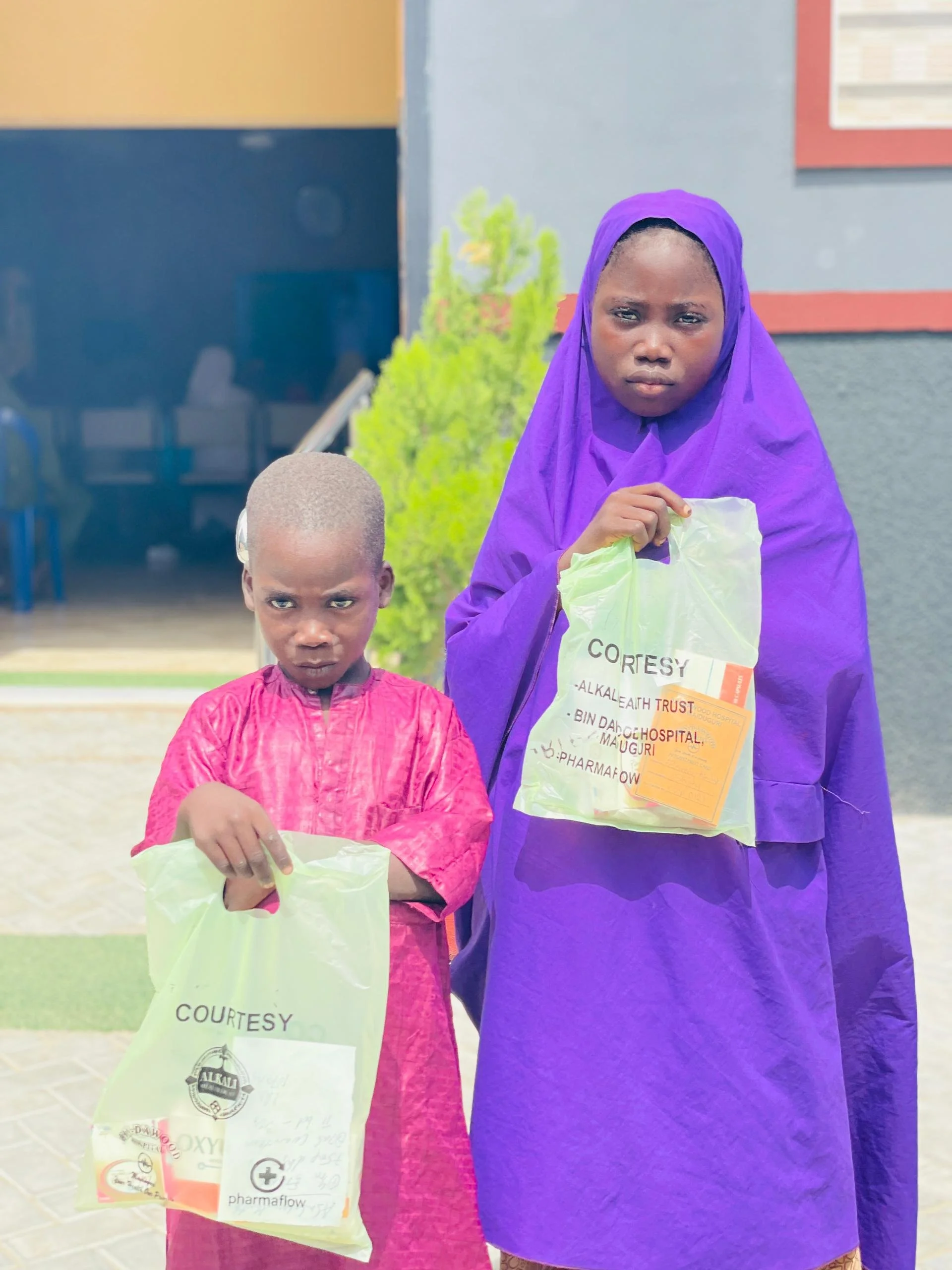 Two children, a girl and a boy, standing outdoors, both holding plastic bags with some documents inside, with serious expressions on their faces.