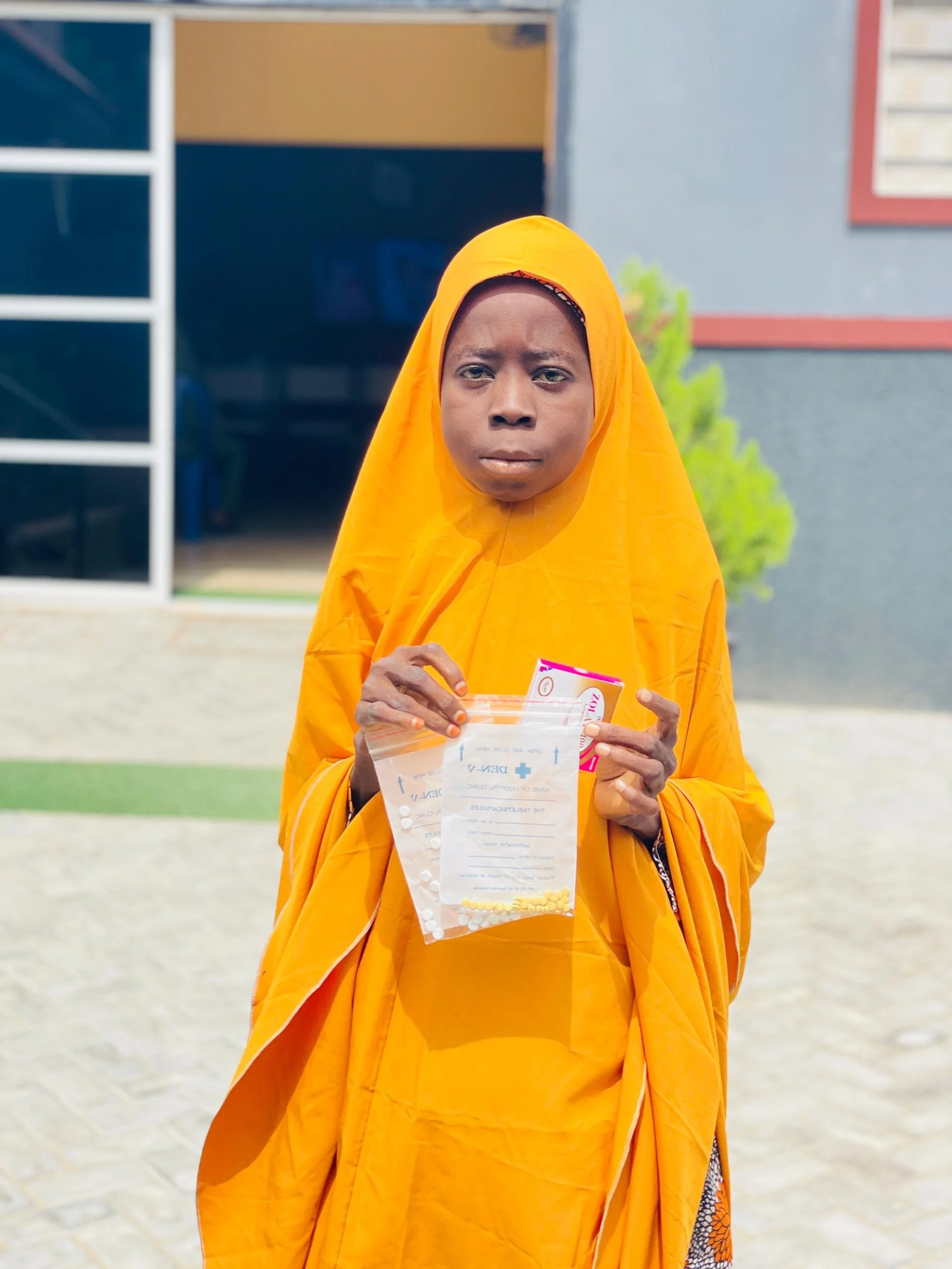 A woman wearing a bright yellow traditional dress and headscarf holding a clear plastic bag with pills and a prescription paper, standing outdoors in front of a building with a gray wall and red window frame.