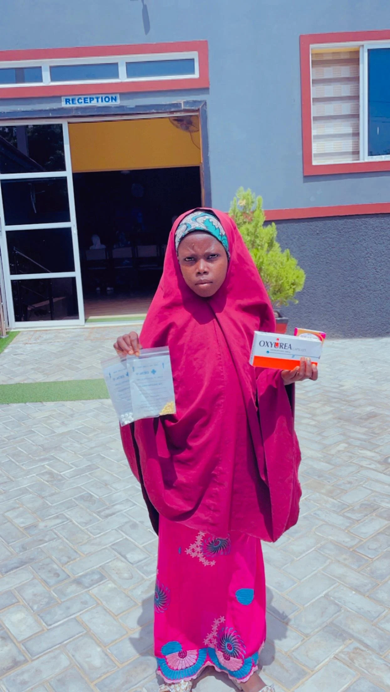 A woman wearing a pink Islamic dress and headscarf standing outside a building labeled 'RECEPTION' holding a box of oxygen and some papers.