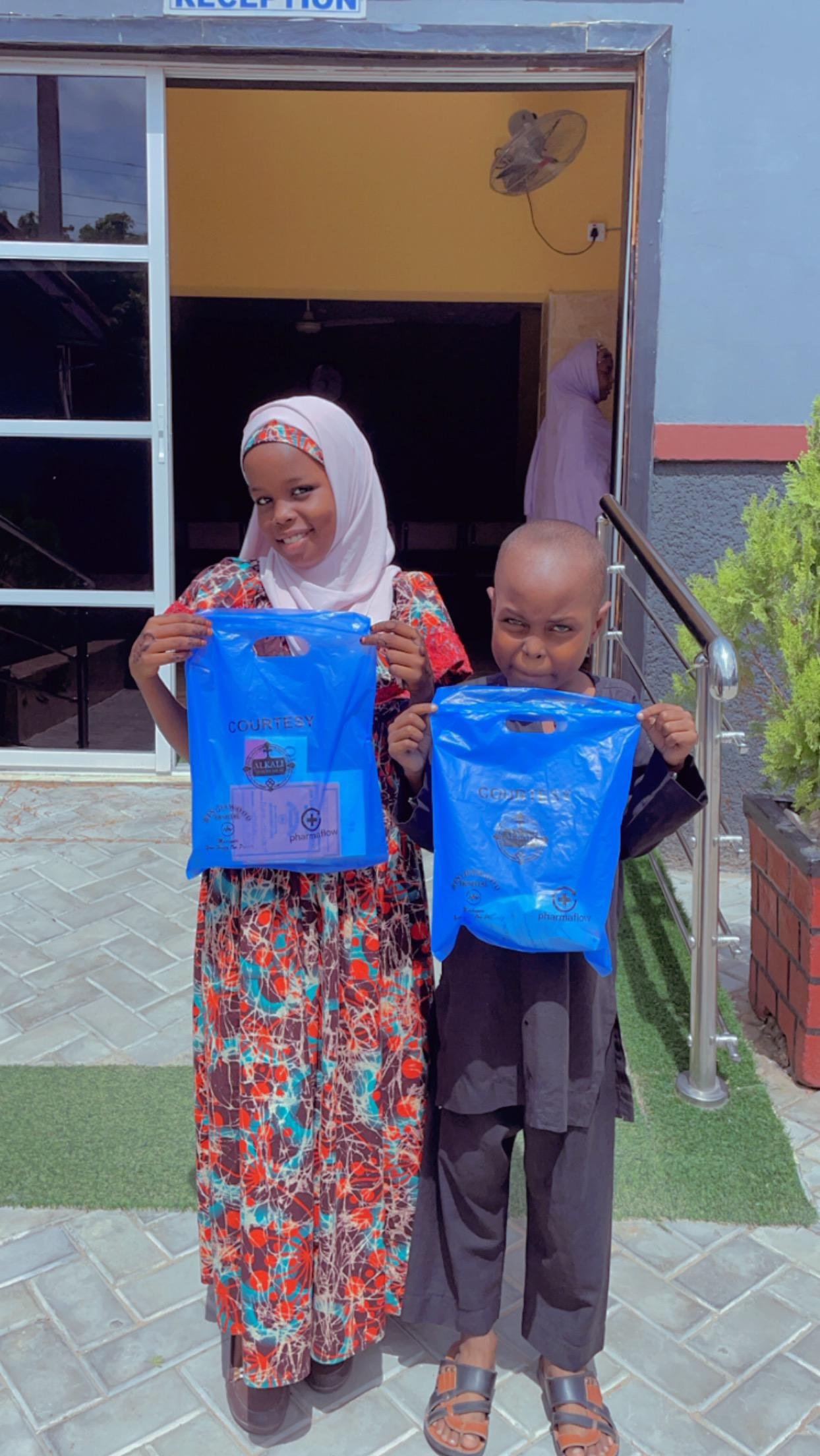 A woman and a young boy standing outside a building, each holding a blue plastic bag, smiling and posing for the photo. The woman is wearing a patterned dress and a white headscarf, while the boy is dressed in black with sandals.