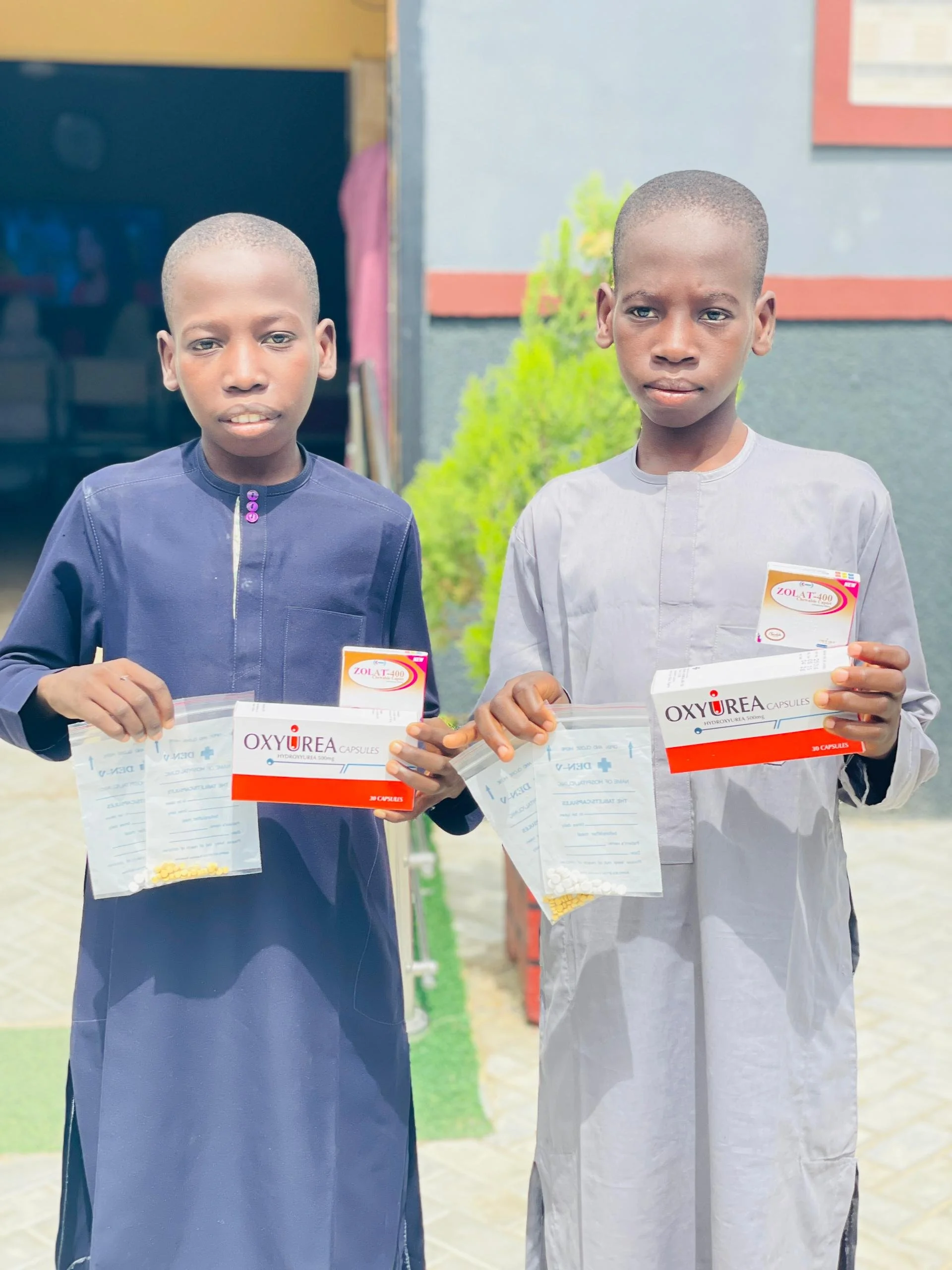 Two young boys standing outdoors holding boxes and packets of medication, looking at the camera.
