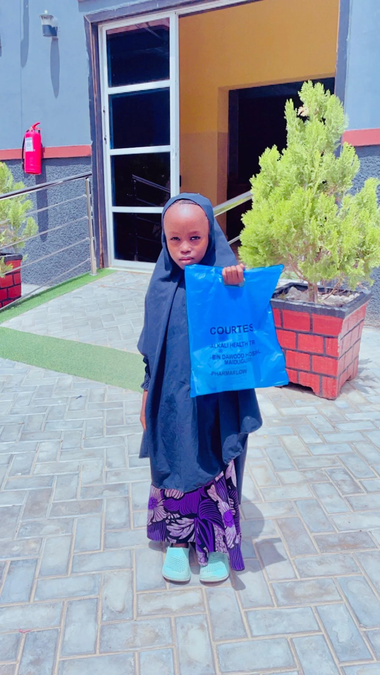 A young girl dressed in traditional clothing and a dark hijab holding a blue shopping bag, standing on a paved outdoor area near a building with plants.