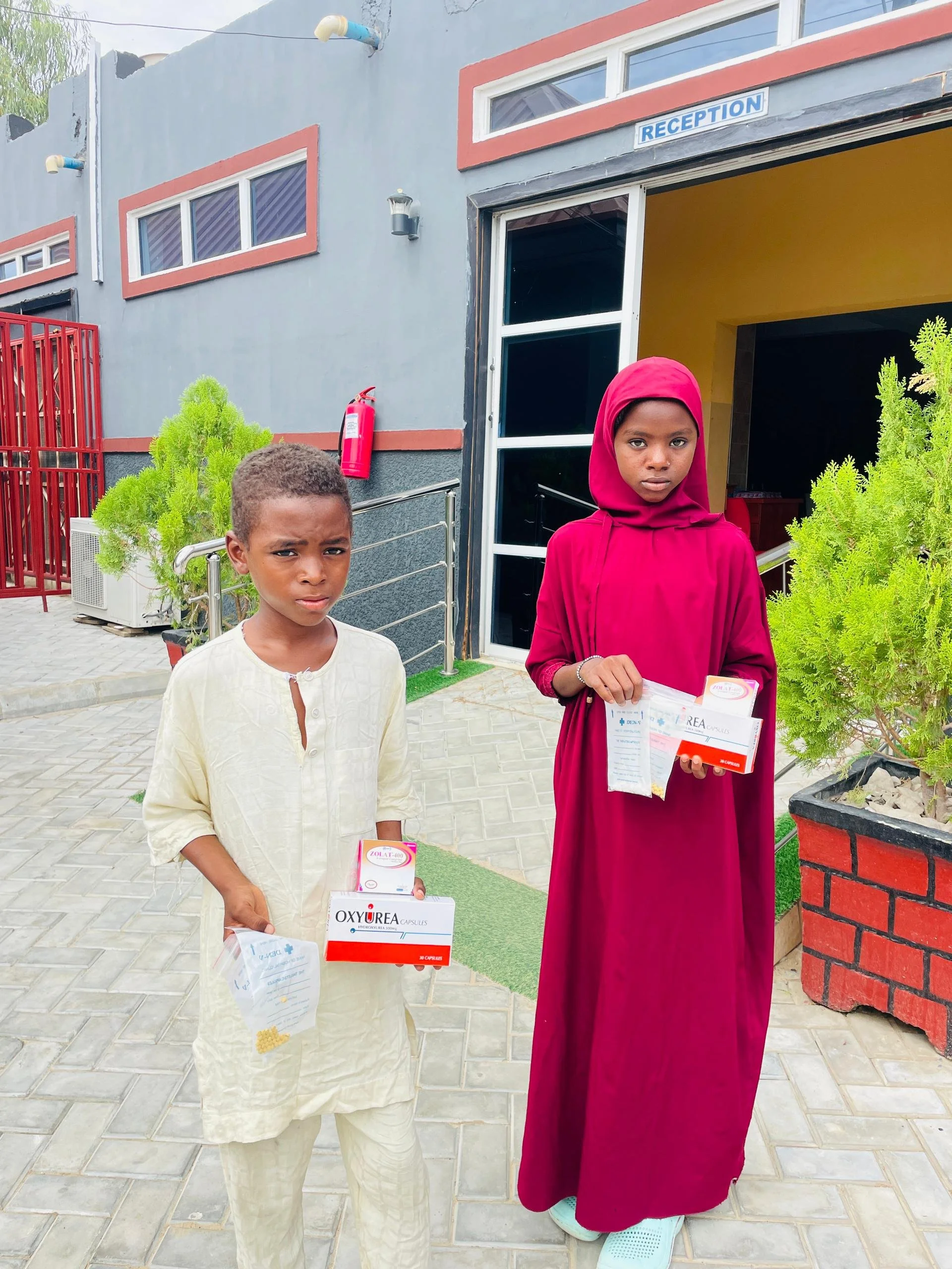 A boy and a girl standing outside a building with a reception sign, each holding medicine boxes and papers.