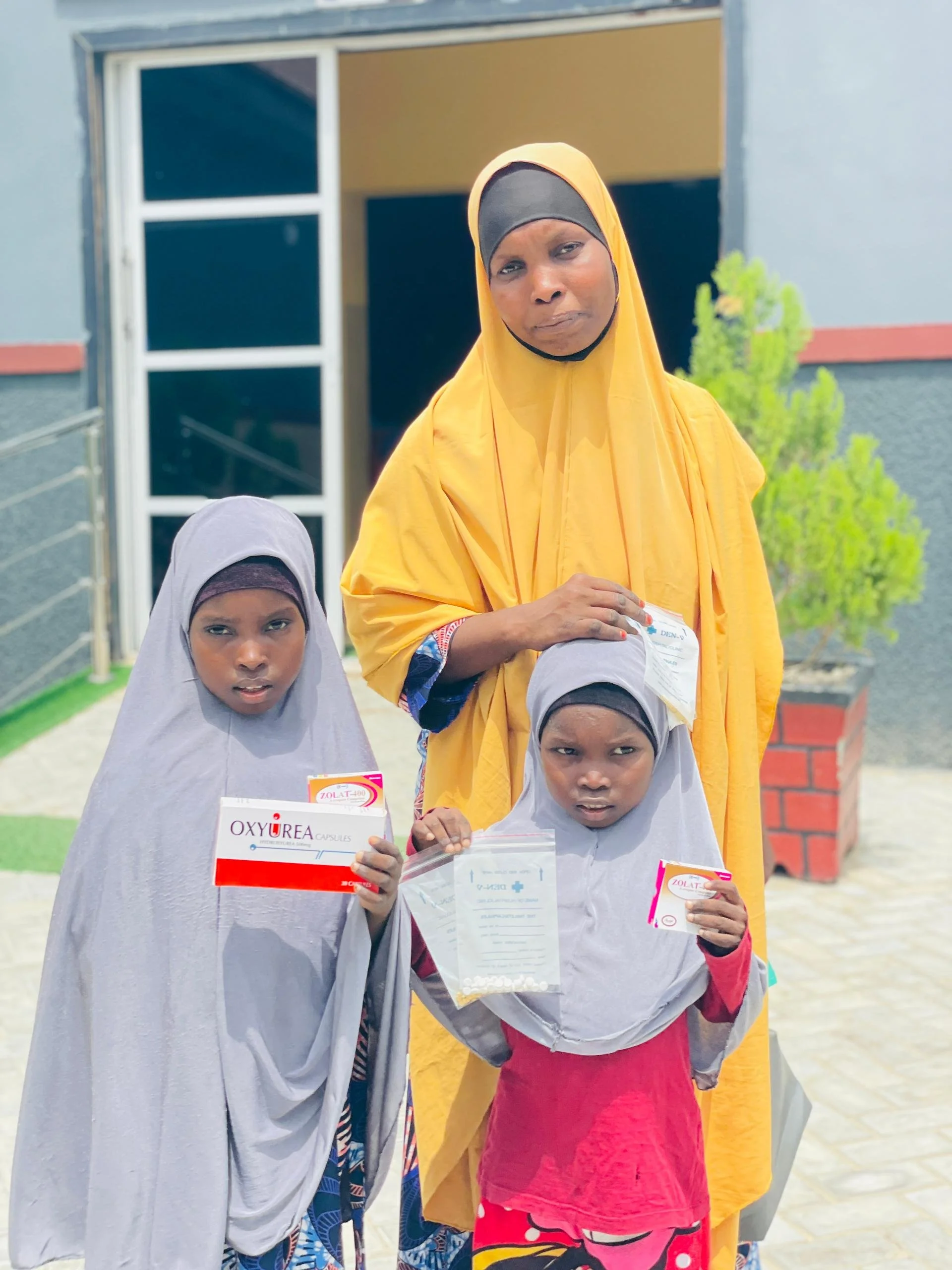 A woman and two young girls standing outdoors in front of a building. The women wears a yellow hijab and the girls wear gray hijabs. The girls hold medication boxes labeled 'OXYUREA' and 'ZOLAT,' and one also holds a document.
