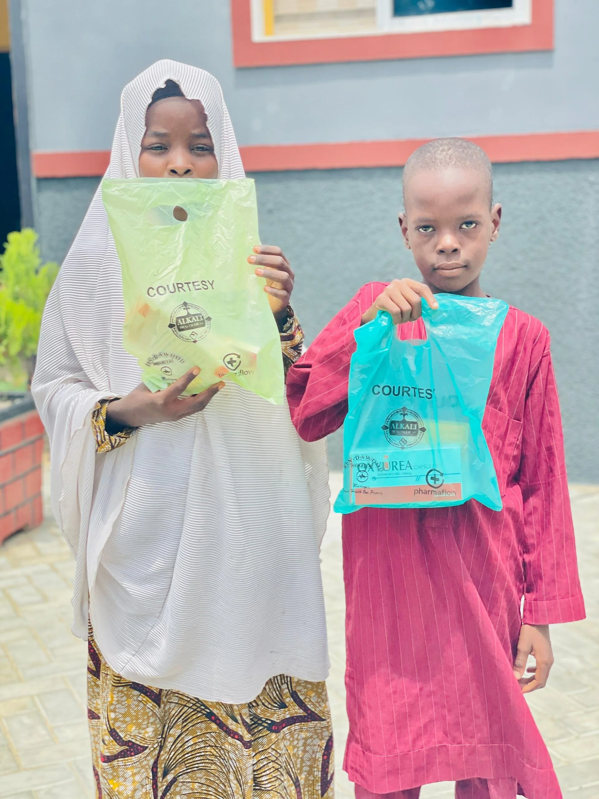 Two children standing outside holding plastic bags with the label 'COURTESY'. The child on the left is wearing a white and black striped head covering and traditional patterned clothing, while the child on the right is dressed in a red striped outfit