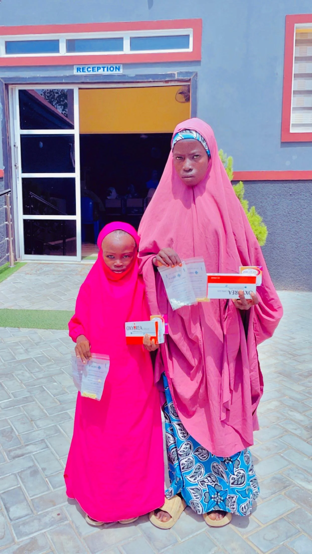 Two women standing outside a building labeled 'Reception' holding boxes of oxygen supply and medical documents.