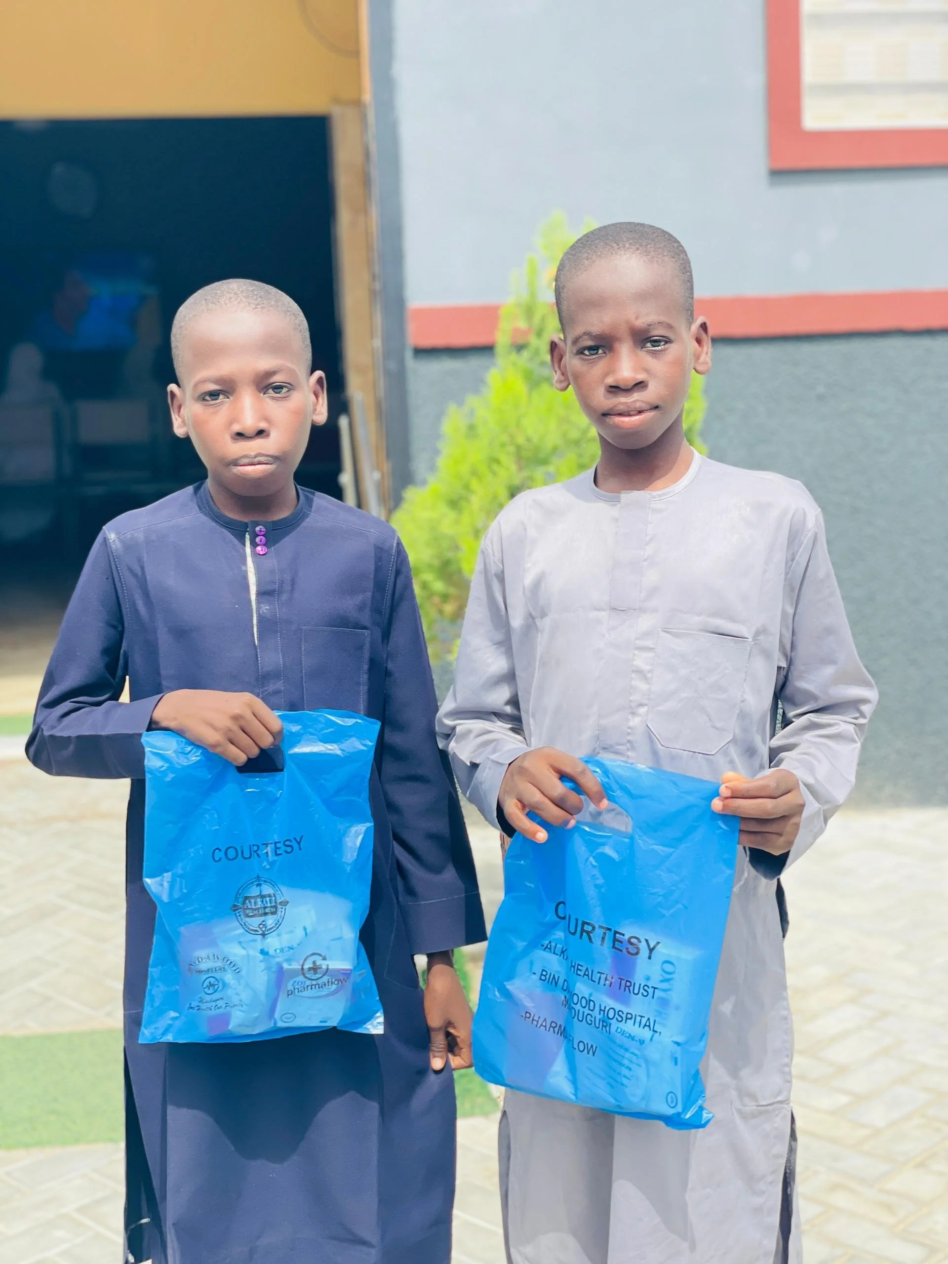 Two boys standing outdoors, each holding a blue plastic bag with the word "COURTESY" printed on it.