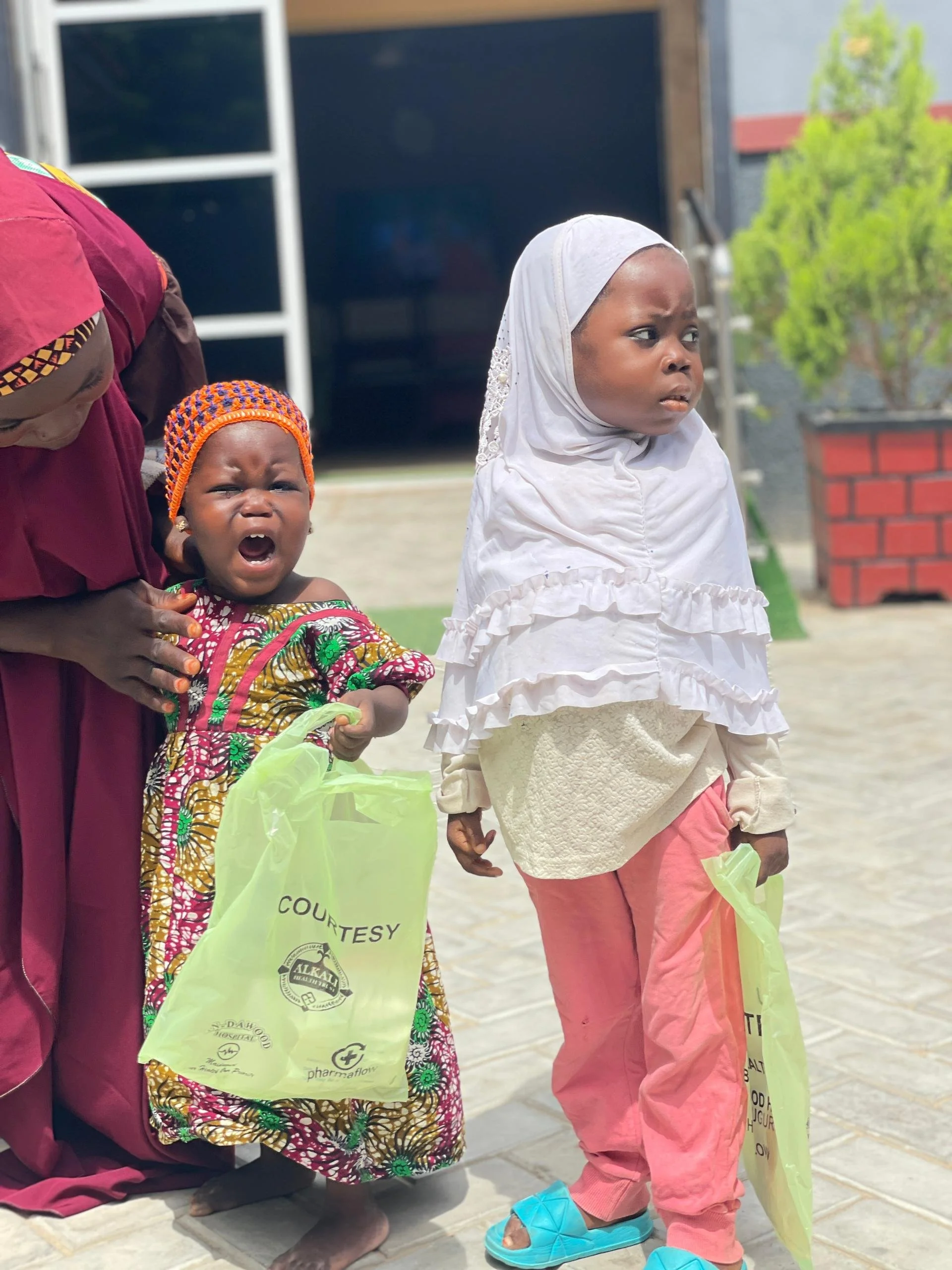 Two young children, a girl and a boy, standing outside on a paved area with a woman partially visible beside them. The girl is wearing a white hooded garment, pink pants, and blue slippers, looking to her right with a serious expression. The boy is w