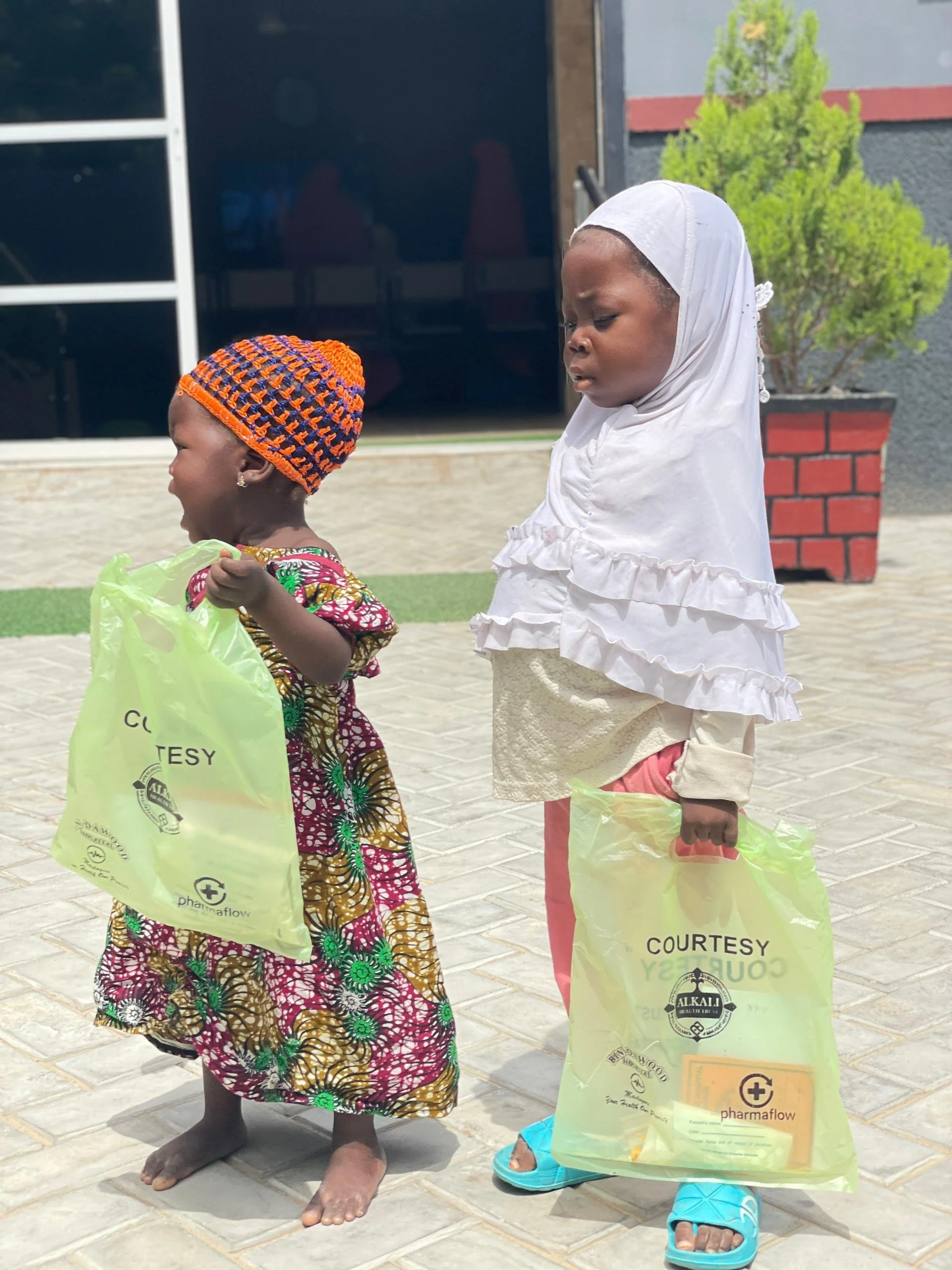 Two young girls standing outside on a tiled surface, each holding a yellow plastic bag with pharmacy branding. One girl wears a colorful dress and a knitted orange and blue hat, smiling and looking away. The other girl wears a white headscarf, light-
