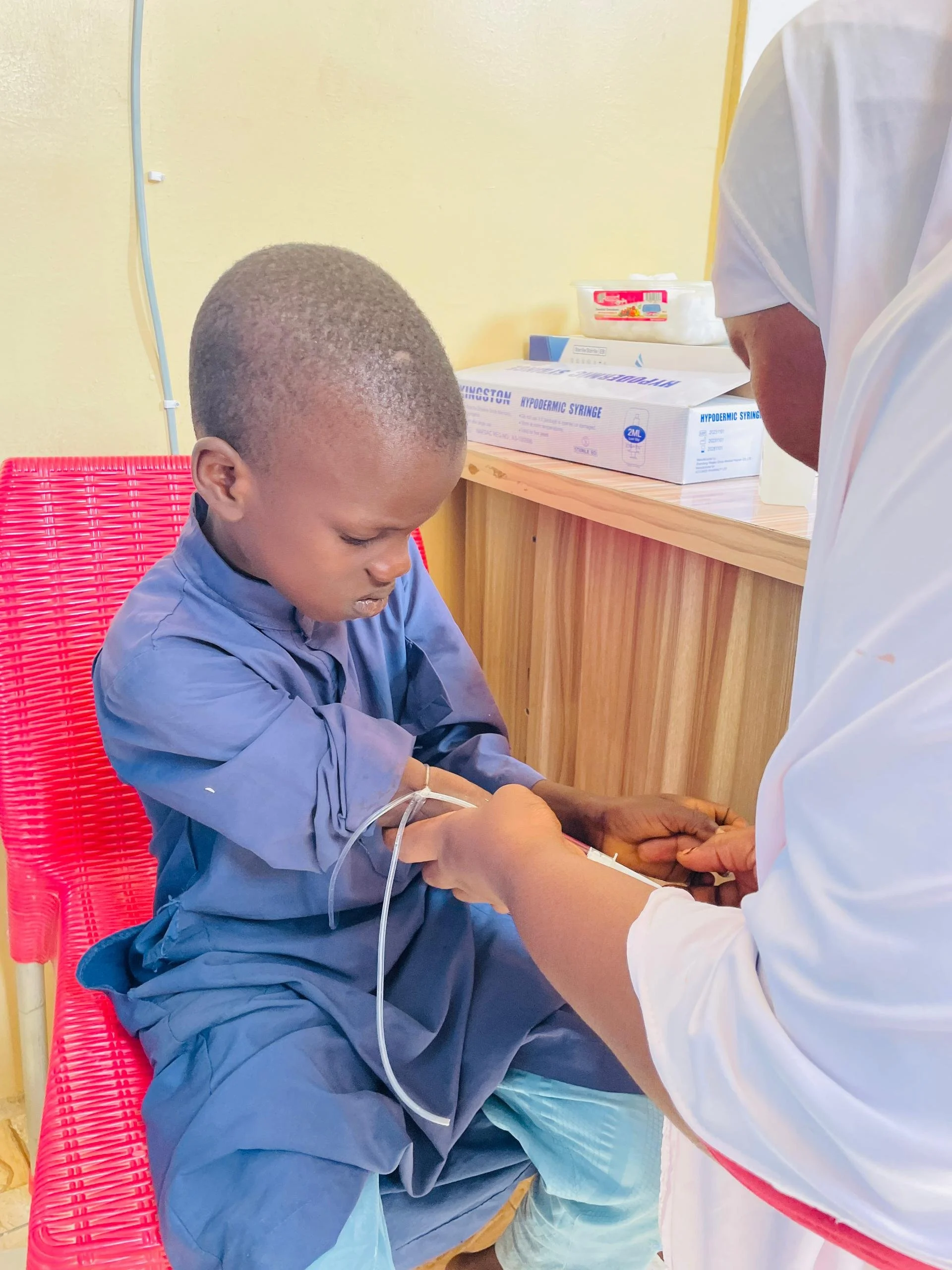 A young boy in a blue hospital gown sits in a red chair, receiving an IV injection from a healthcare professional dressed in white. The scene is inside a medical clinic with yellow walls and medical supplies on a wooden counter in the background.