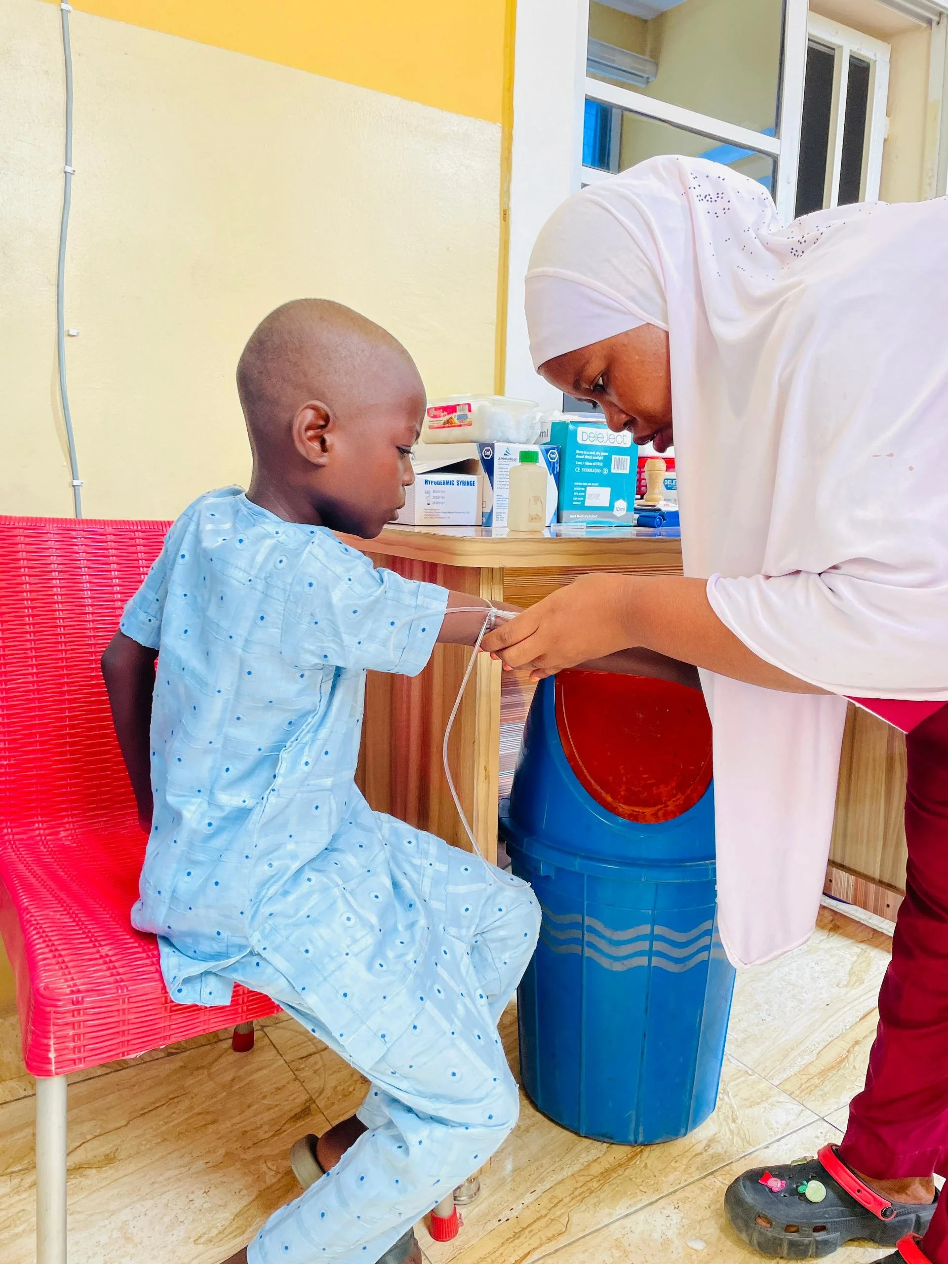 A young boy in hospital gown sitting on a pink chair with a IV in his arm, receiving medical treatment from a woman dressed in white, possibly a nurse, in a brightly lit medical facility.