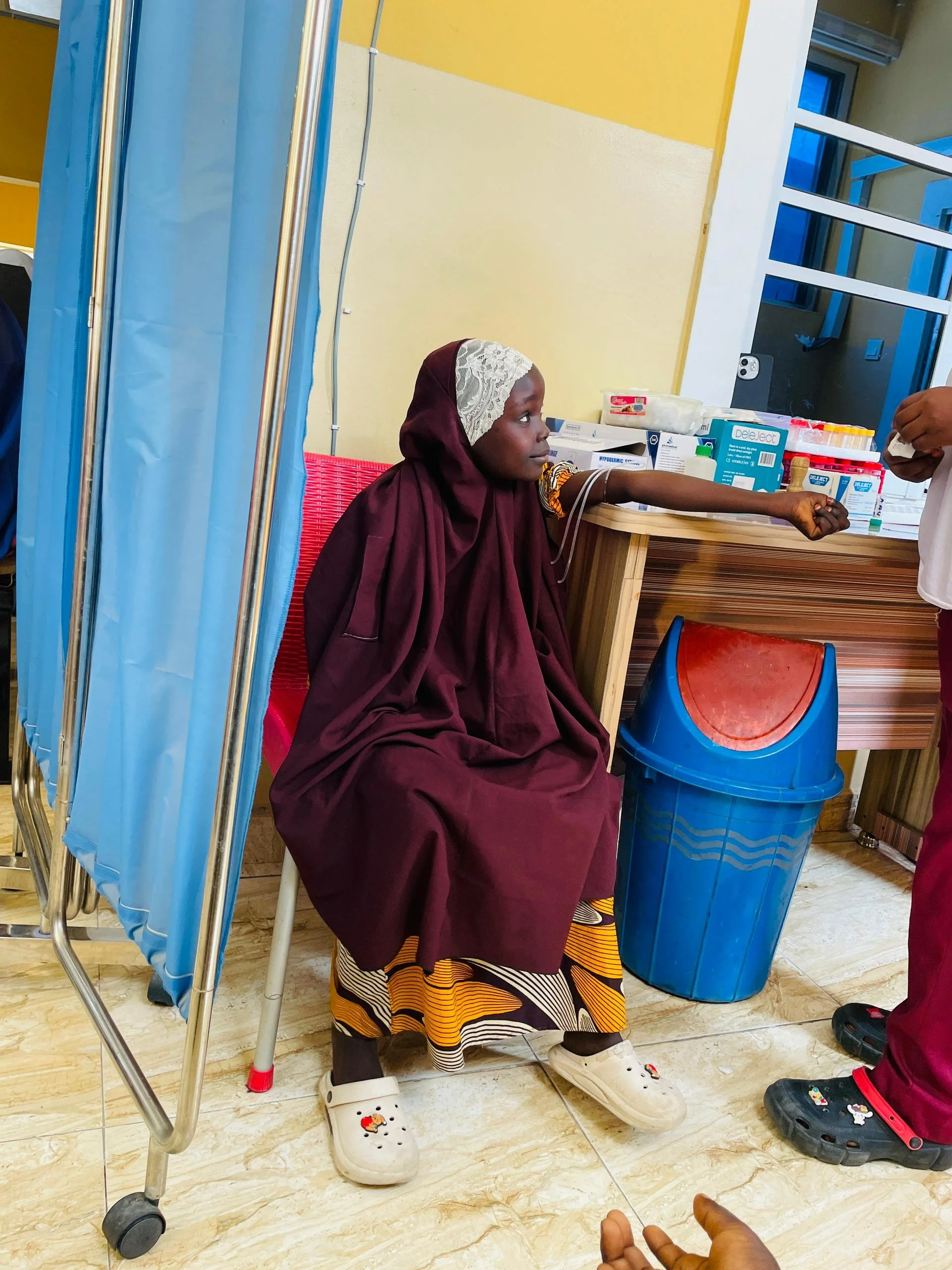 A girl sitting on a bench with her arm extended, receiving a medical injection in a clinic. She is wearing traditional clothing and white Crocs with a cartoon character. There are medical supplies and a trash bin behind her.