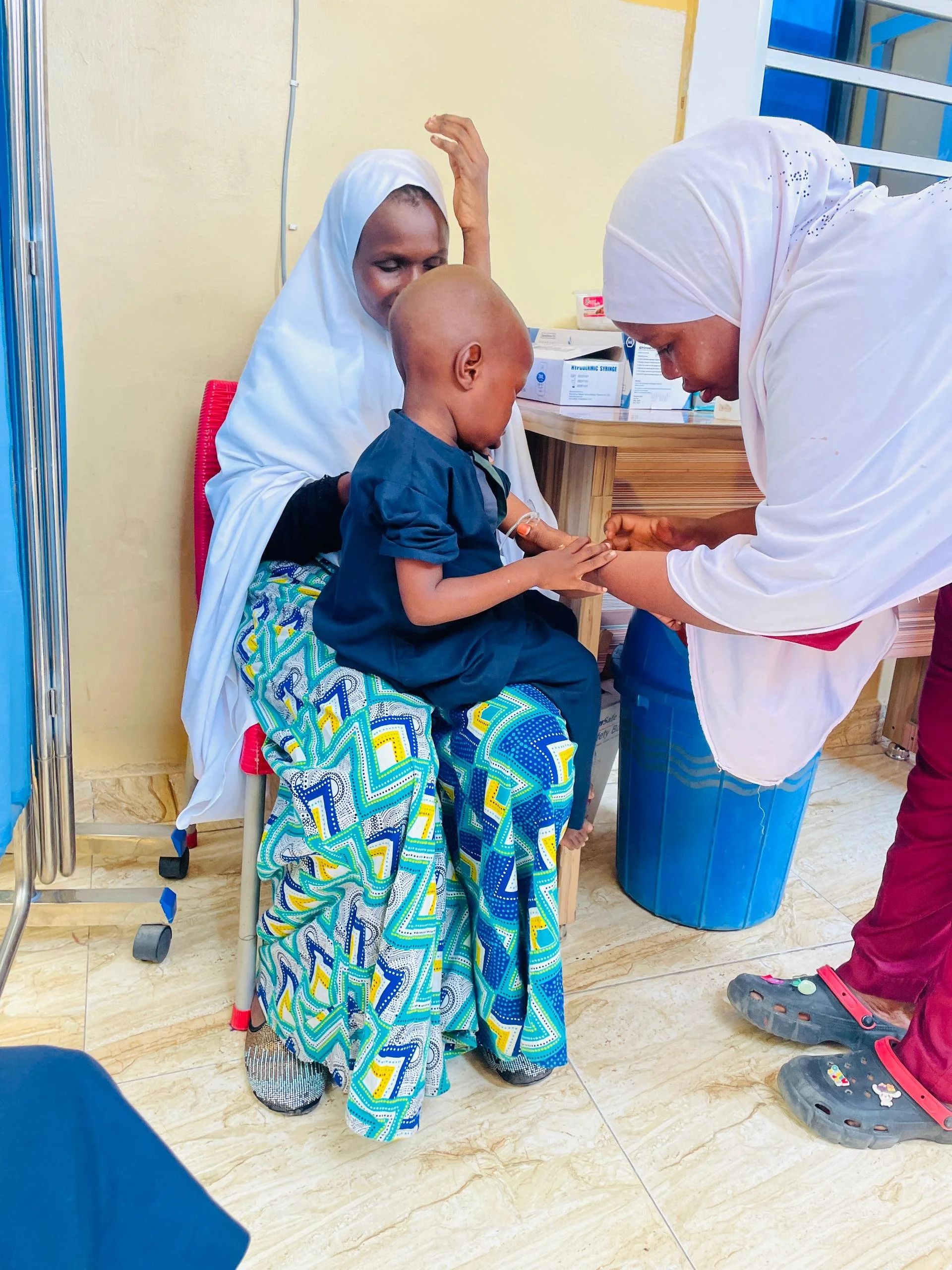 A young boy receiving an injection from a nurse while seated on a woman's lap at a medical clinic.